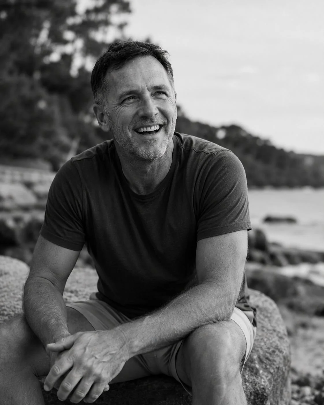 A smiling man with gray hair and a beard, wearing a dark t-shirt, sitting on a rock at the beach, in black and white.