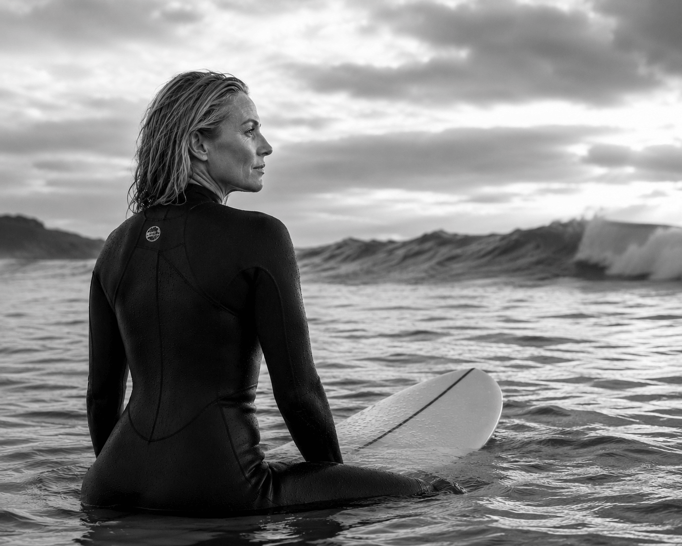 A woman in a wetsuit sitting on a surfboard in the ocean, looking out at the waves with mountains in the distance under a cloudy sky.