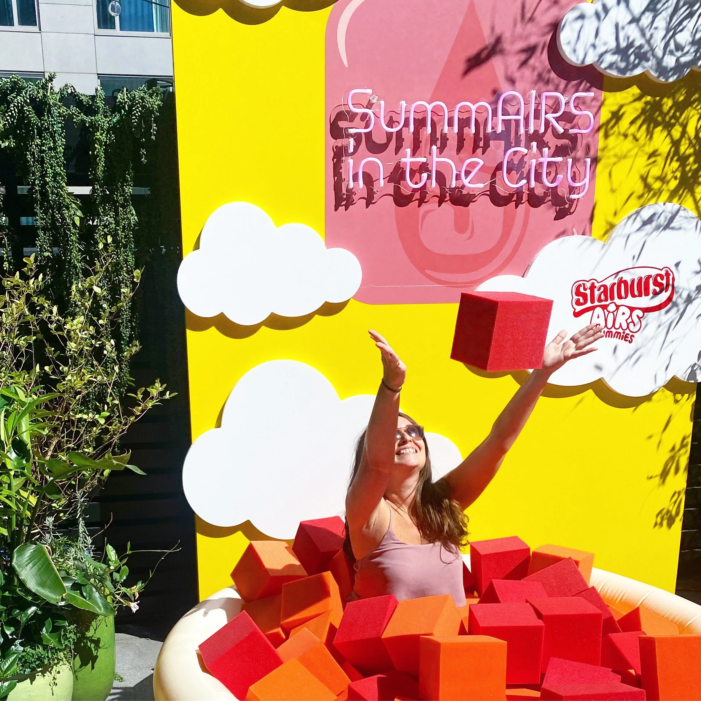 A woman with sunglasses smiling, reaching up with both hands, standing behind large red and orange foam blocks arranged as part of a summer-themed display. The background includes a bright yellow panel with white cloud shapes, a pink rectangle with purple neon text, and some greenery on the left side.