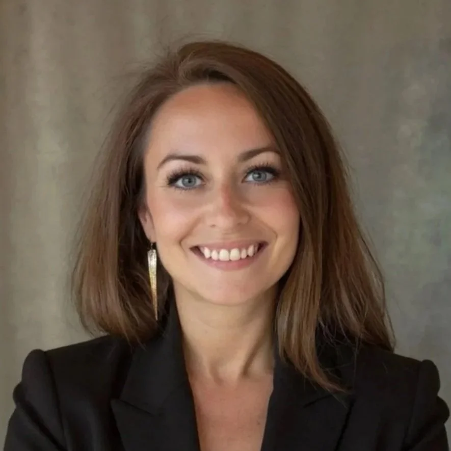 A woman with shoulder-length brown hair, blue eyes, and light skin, smiling and wearing a black blazer and gold earrings, against a neutral background.