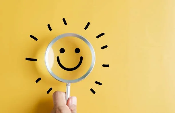 Mano che tiene una lente con un volto sorridente disegnato, circondato da linee che rappresentano i raggi del sole su sfondo giallo.