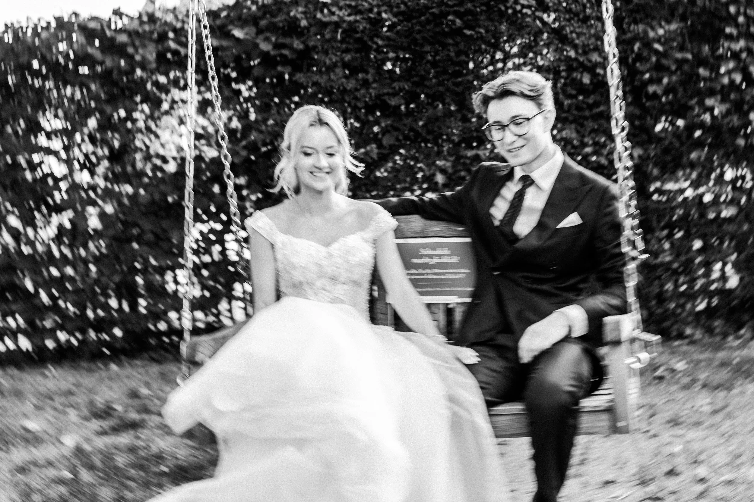 Ein lachendes Paar sitzt auf einer Schaukel im Freien, umgeben von Bäumen, und trägt formelle Kleidung, vermutlich bei einer Hochzeit.