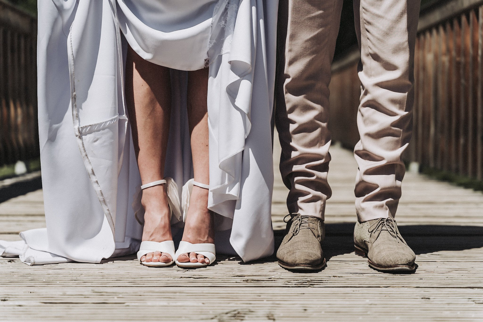 Nahaufnahme der Beine zweier Personen, eine Person trägt ein weißes Kleid und Sandalen, die andere Person trägt beige Hosen und braune Schuhe, auf einem Holzsteg.