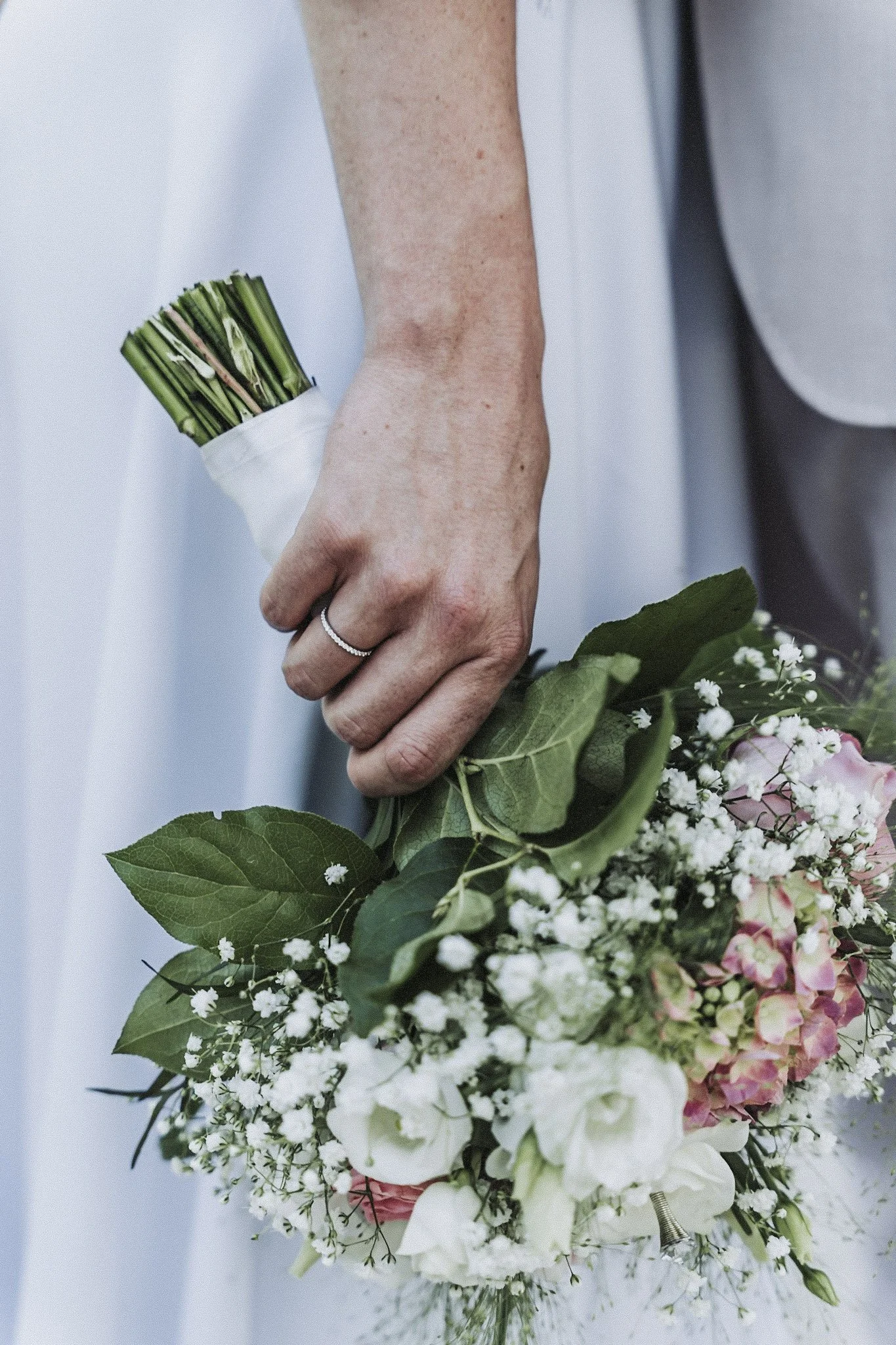 Nahaufnahme einer Hand, die eine Hochzeitsblume hält, bestehend aus weißen und rosa Blumen mit grünen Blättern.