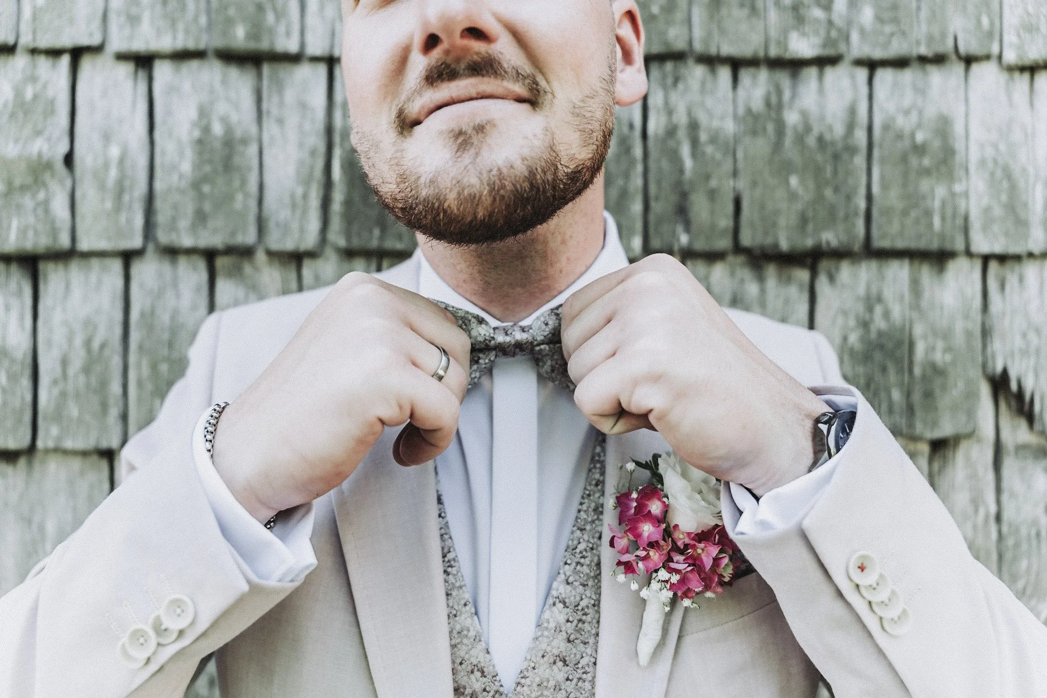 Nahaufnahme eines Mannes in formeller Kleidung, der seinen Krawattenknoten festzieht. Er trägt eine weiße Jacke mit Blumen im Buttonhole und steht vor einer Holzwand.