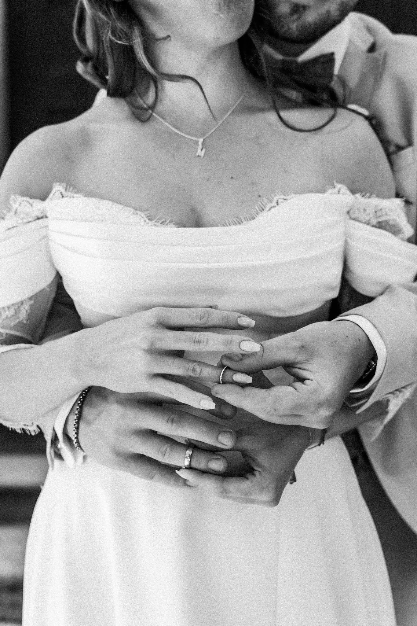 Schwarz-Weiß-Fotografie eines Paares bei einer Hochzeit, Nahaufnahme ihrer Hände, die sich um die Taille des Brautpaares legen, Braut trägt ein off-shoulder Kleid und eine Hochzeitskette.