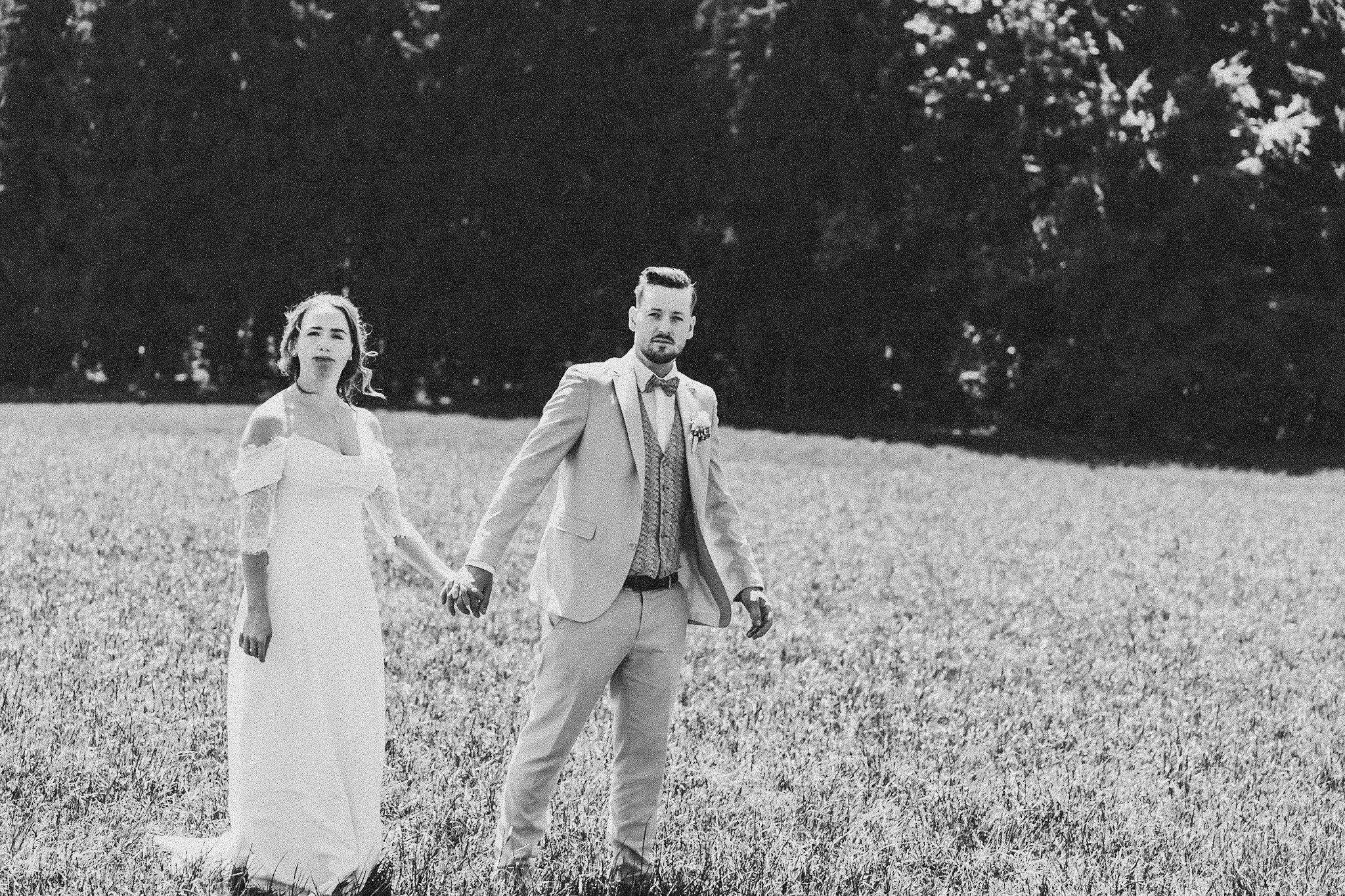 Ein Brautpaar, ein Mann und eine Frau, hält Händchen im Freien auf einer Wiese, umgeben von Bäumen, schwarz-weiß Foto.