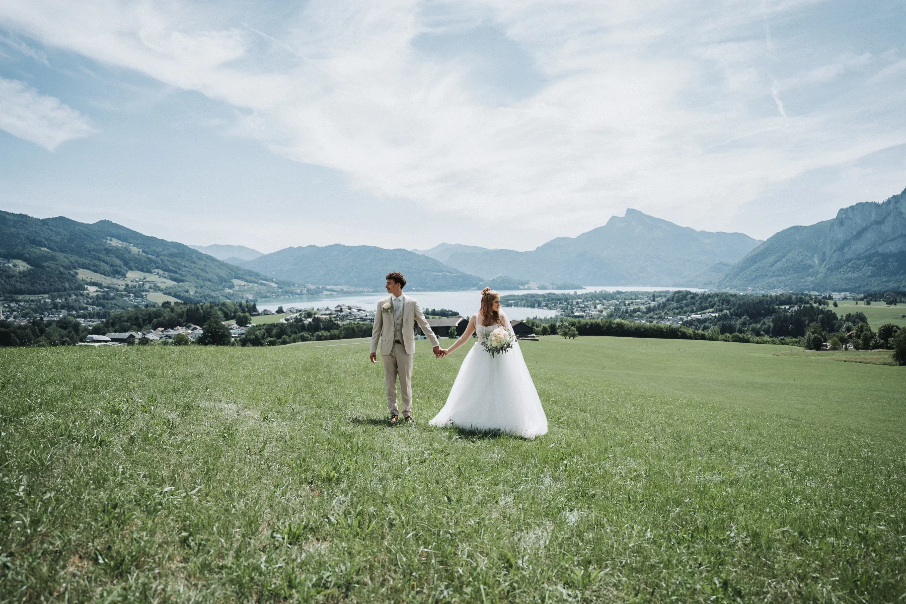 Brautpaar auf einer Wiese mit See und Bergen im Hintergrund.