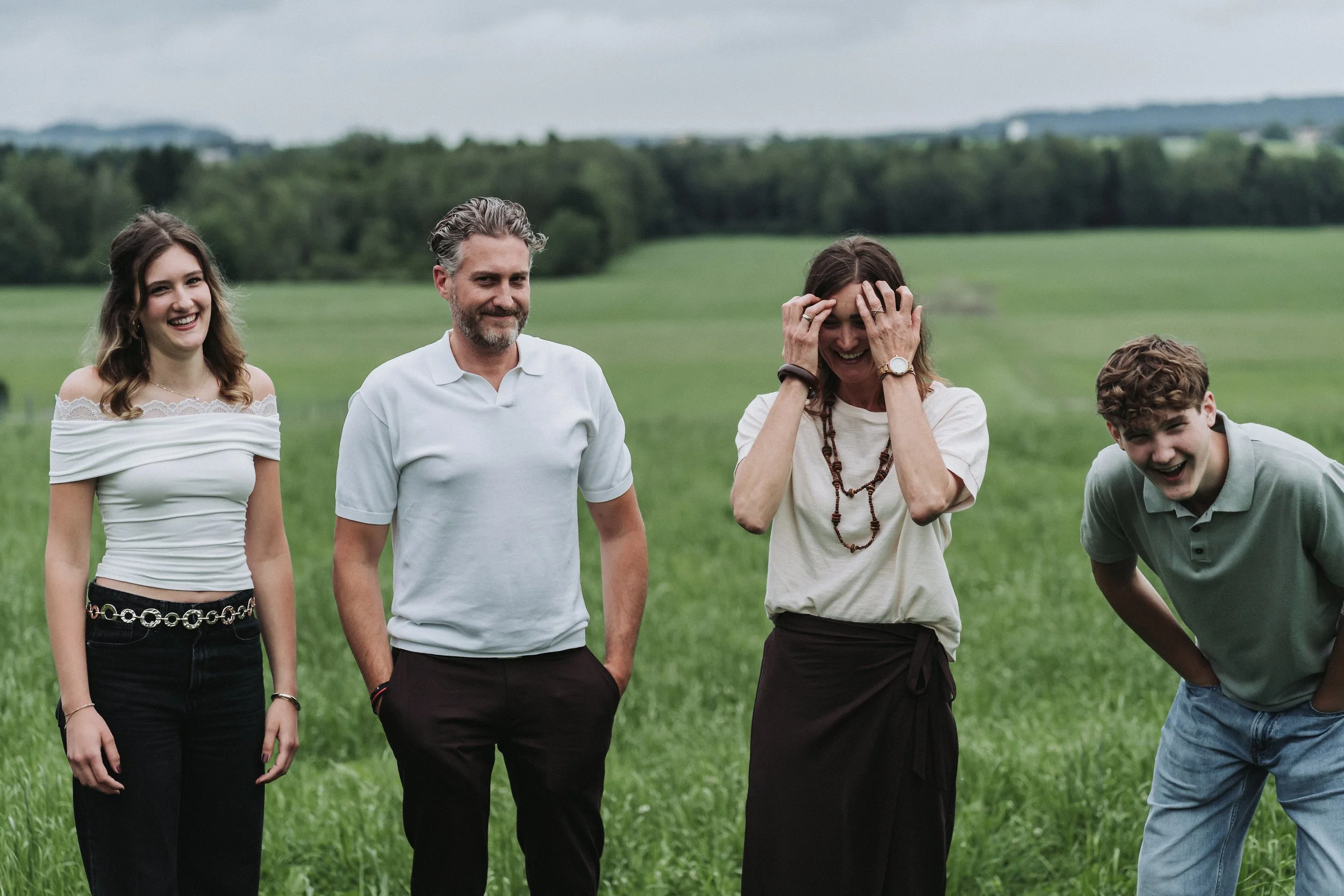 Fünf lachende Menschen, drei Frauen und zwei Männer, stehen in einem grünen Feld bei bewölktem Himmel, sind entspannt und genießen die Gesellschaft.