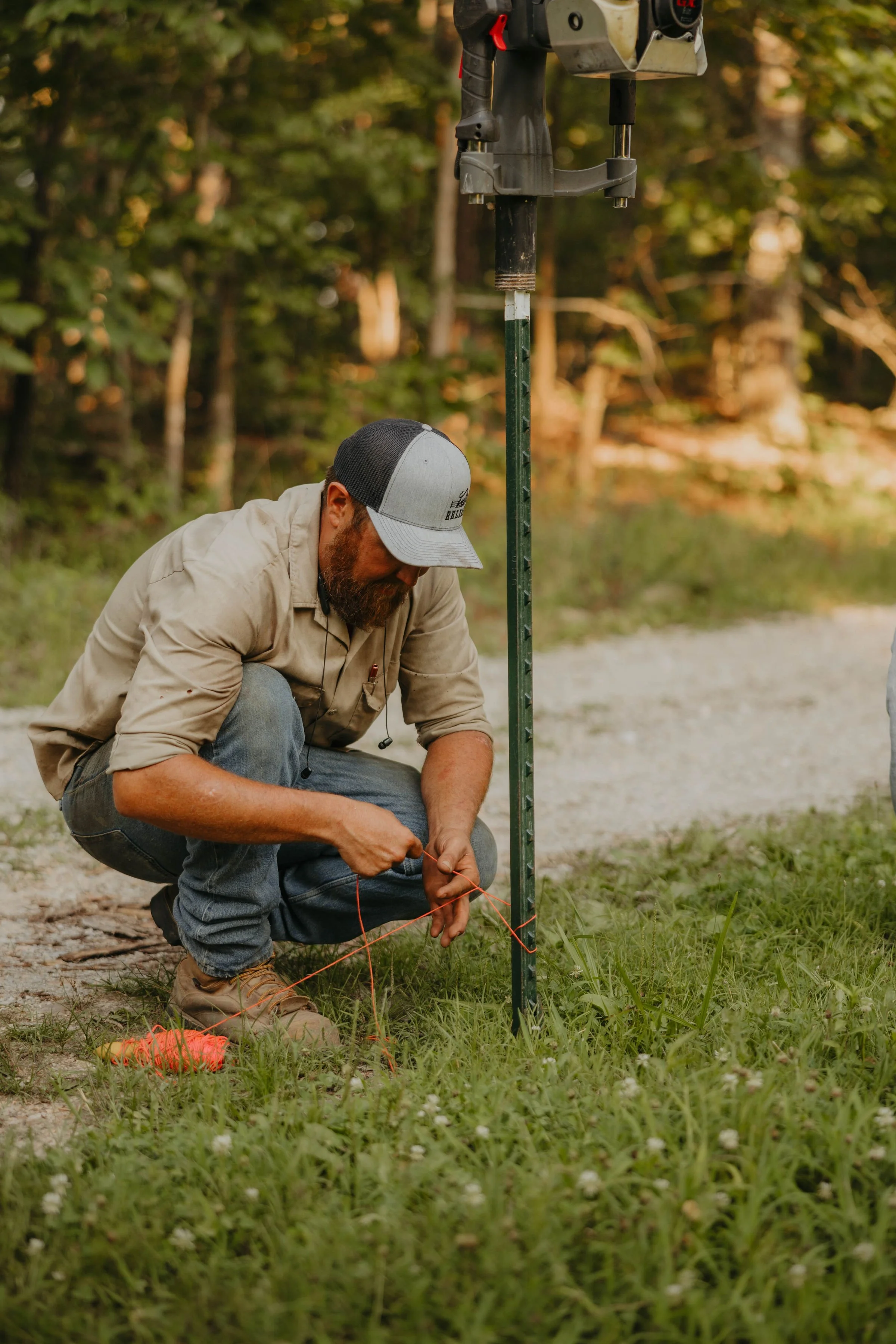 A person squatting on the grass outdoors, working with red string, near a metal pole with a mounted device, in a wooded area during daylight.