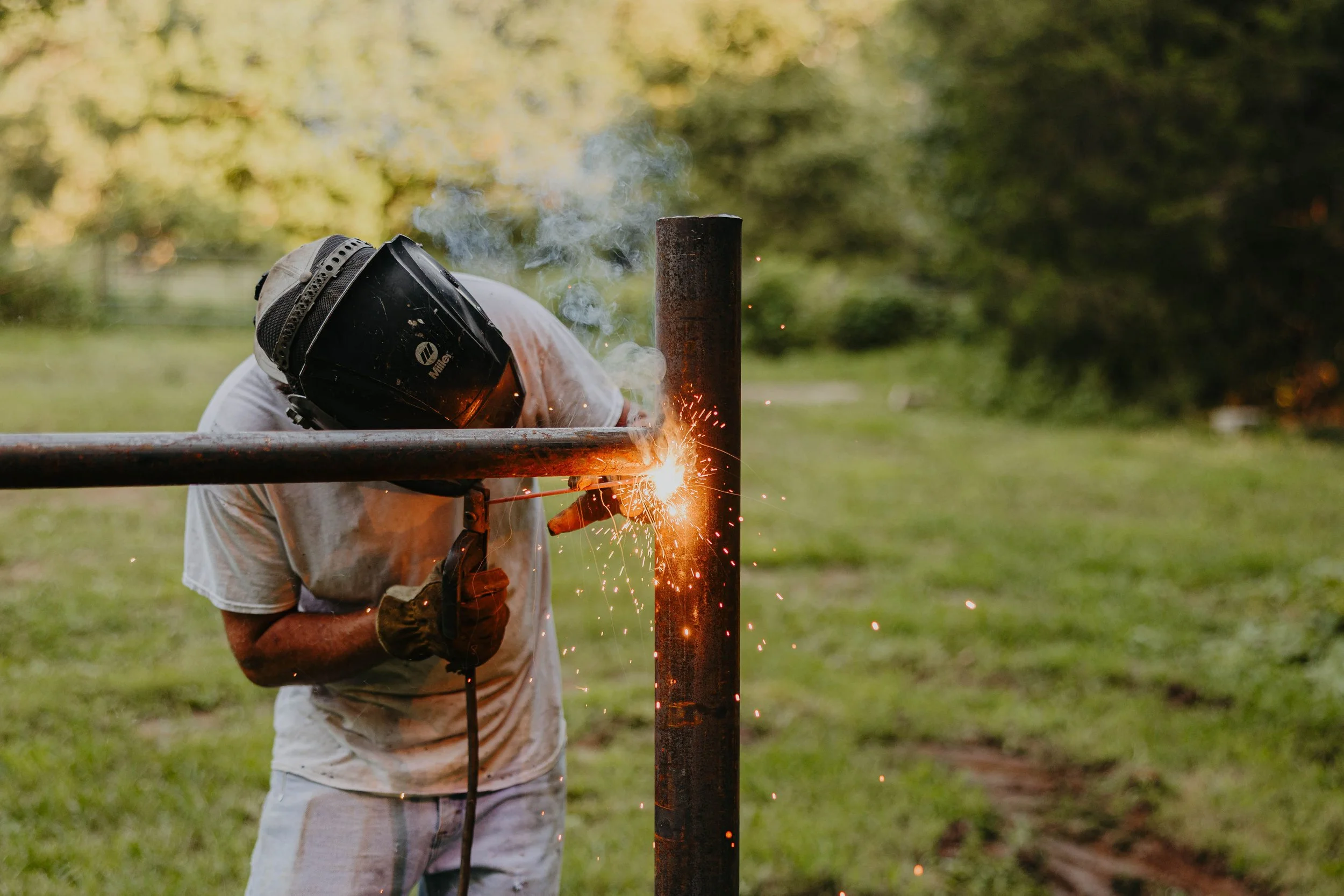 A person wearing a welding helmet and gloves welding a metal pipe fence outdoors, with sparks and smoke visible around the welding area.