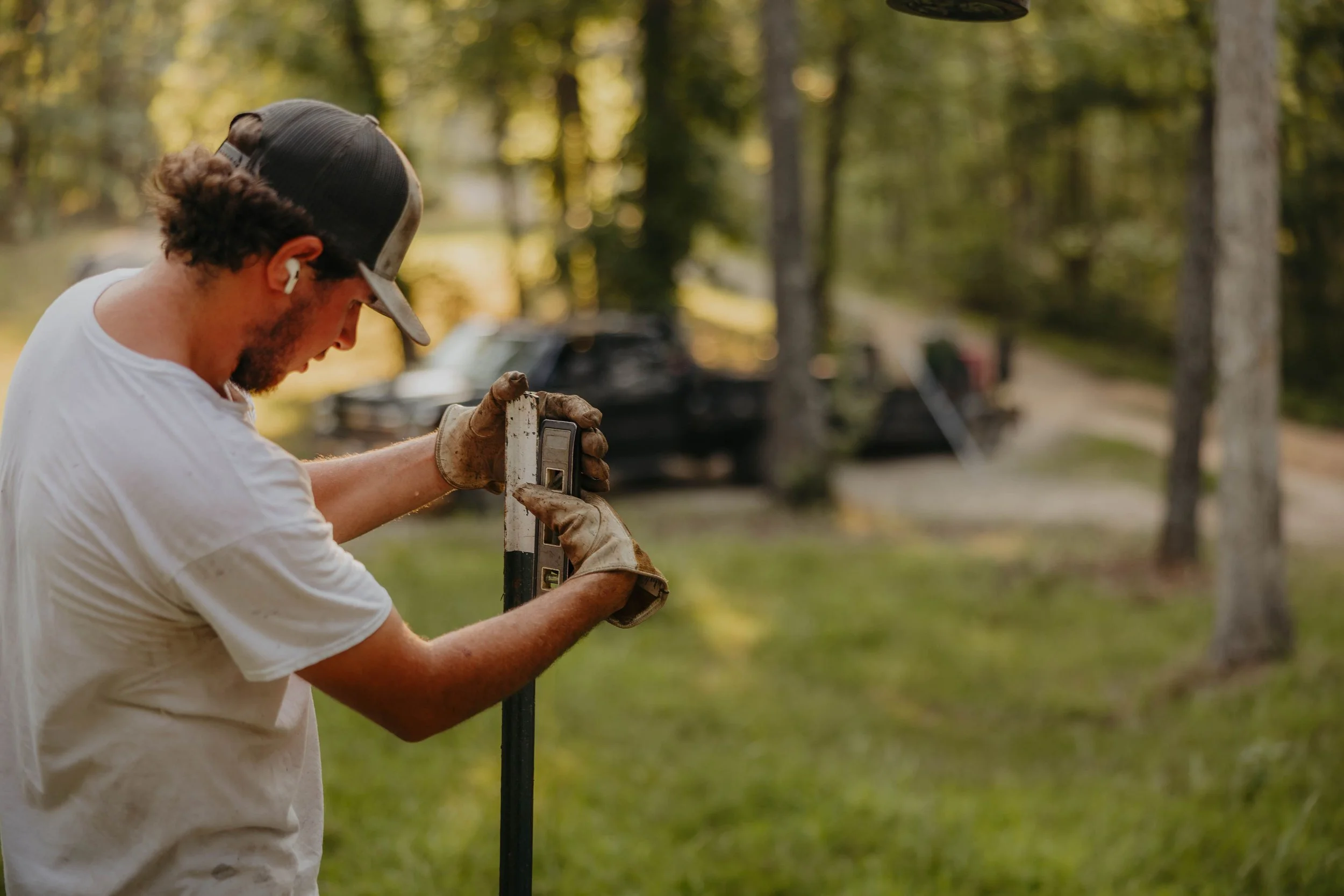 A man wearing a baseball cap, white T-shirt, and gloves using a level tool outdoors in a wooded area with trees and blurred vehicles in the background.