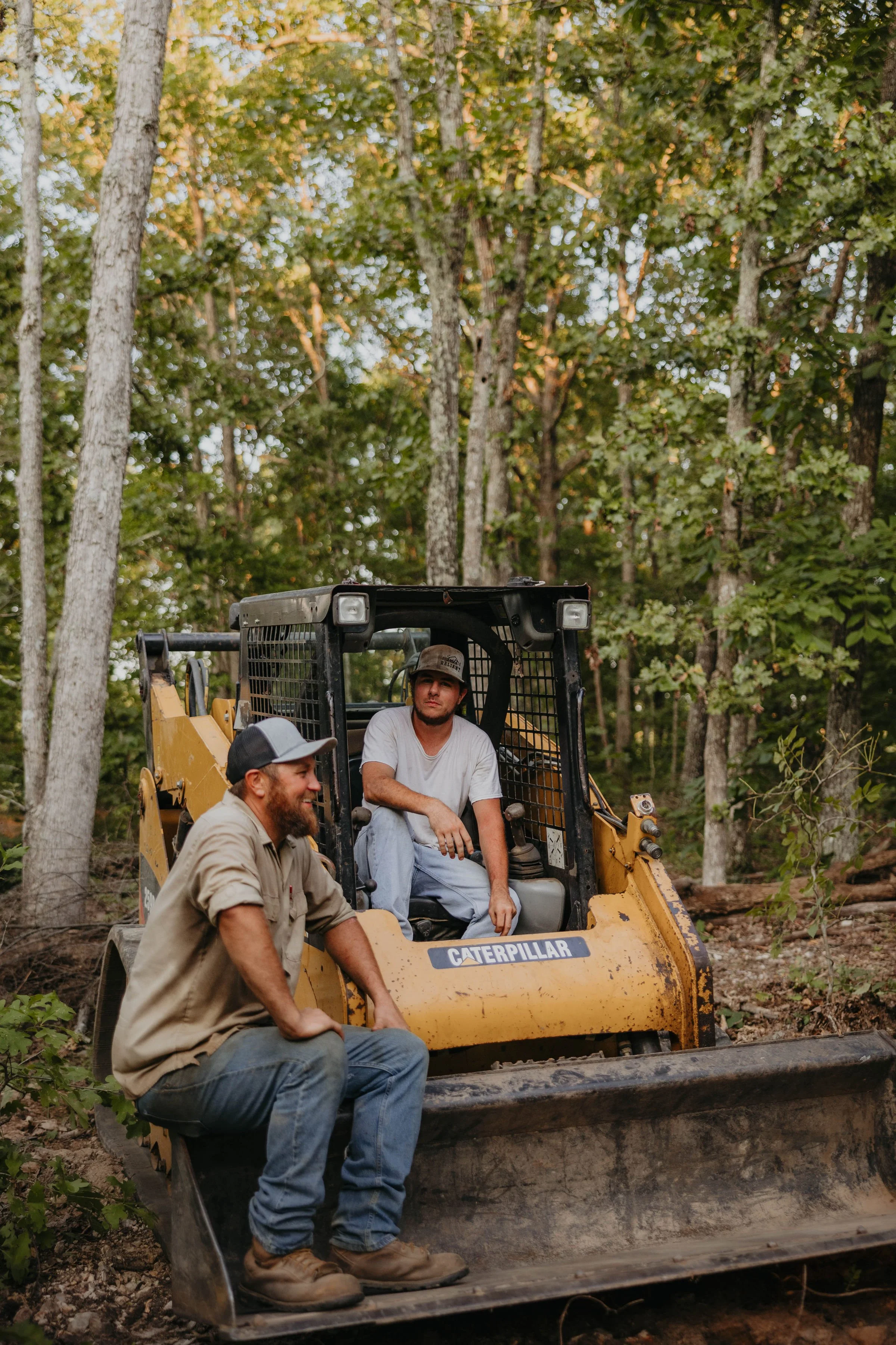 Three men are sitting on a yellow Caterpillar bulldozer in a wooded area with tall trees and green foliage. Two men are sitting inside the bulldozer, one with a beard and wearing a cap and a beige shirt, the other with a beard and wearing a light gray shirt. The third man is kneeling in front of the bulldozer, wearing a cap and a beige shirt. The ground is dirt and the scene appears to be outdoors in a forested setting.