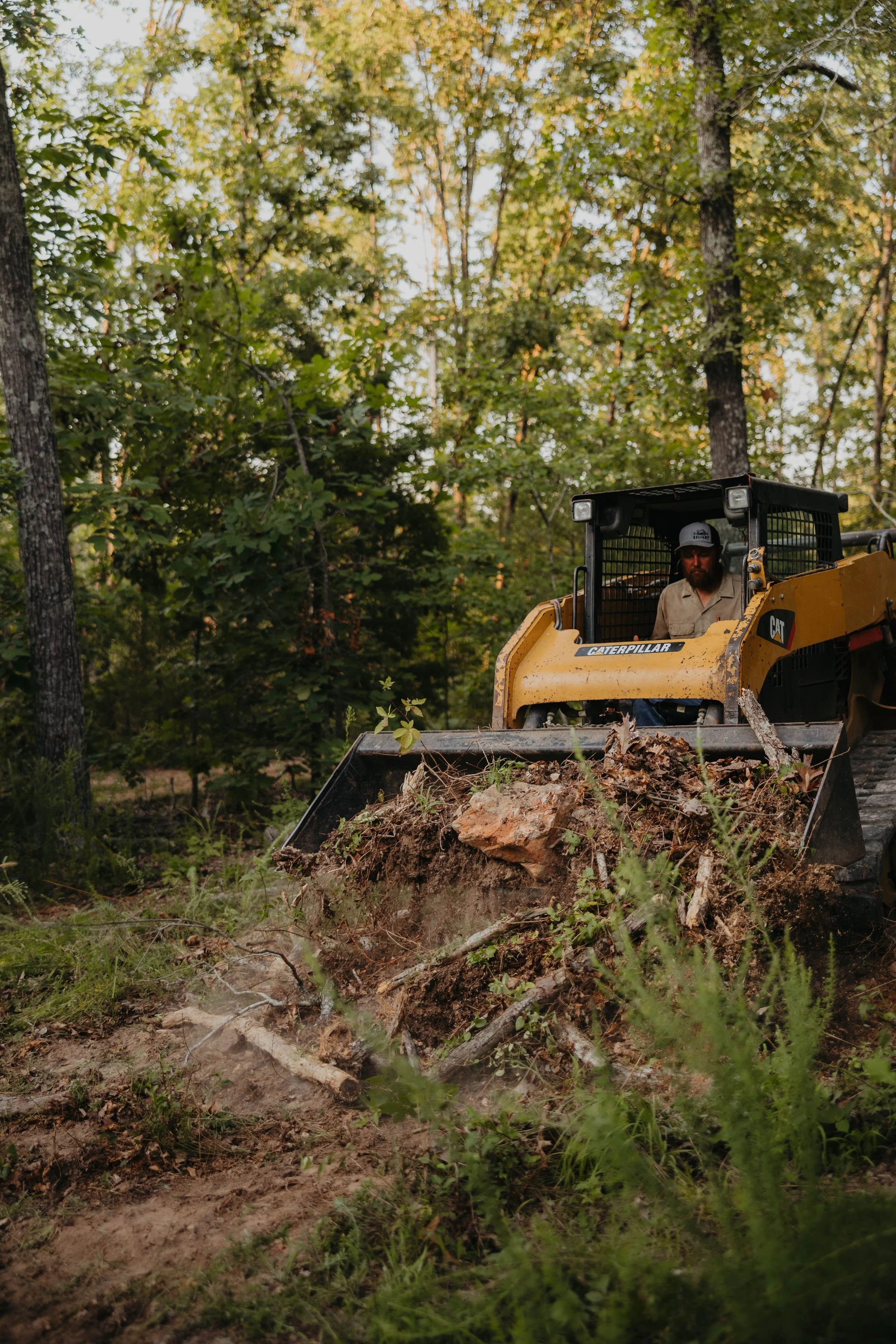 A man operating a yellow Caterpillar bulldozer in a forest, pushing dirt and debris, with trees and green foliage in the background.