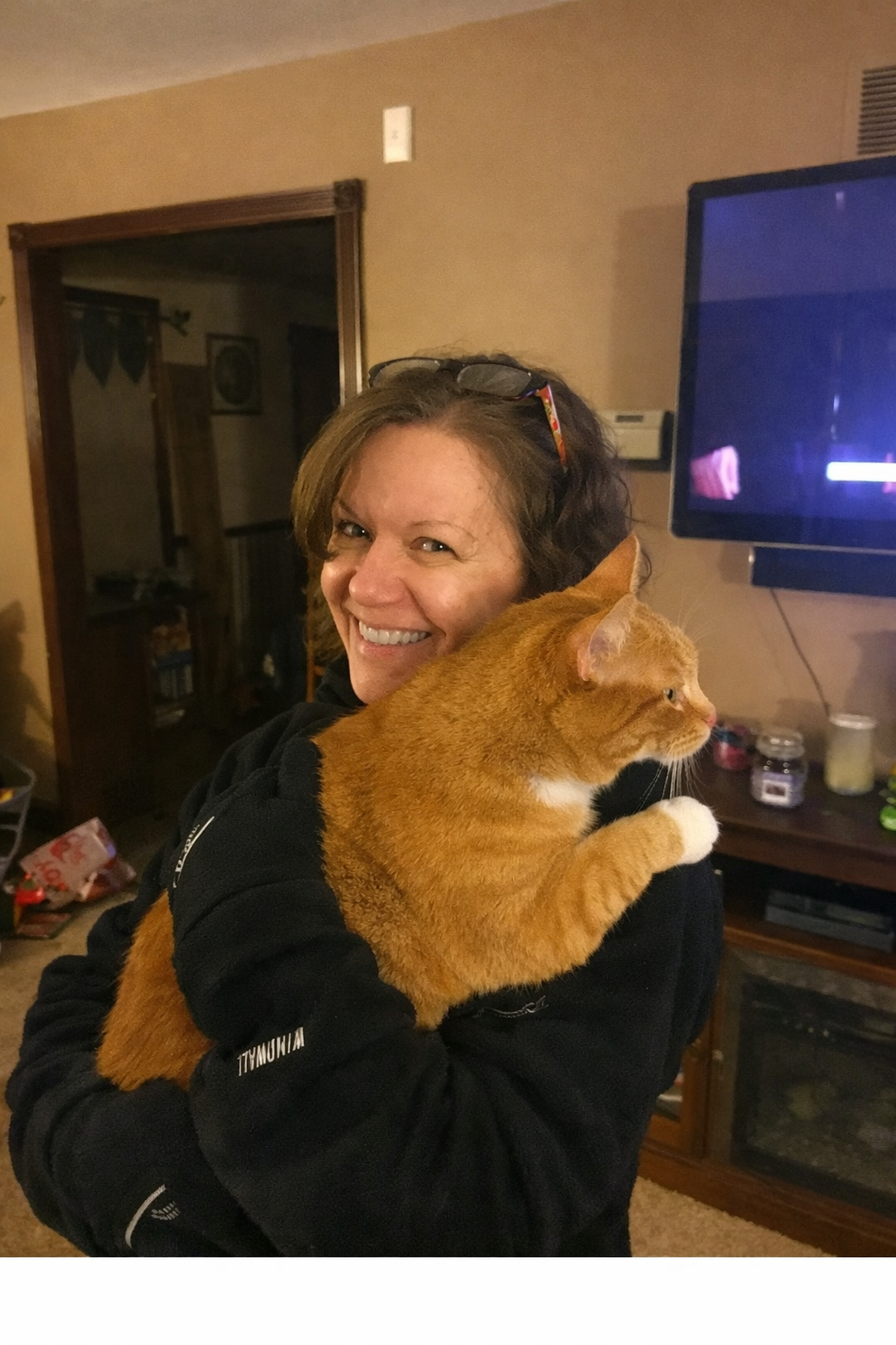 A woman smiling while holding an orange tabby cat inside a home.
