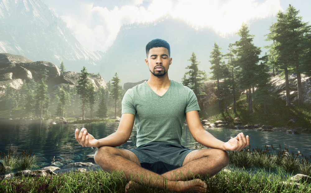 A man meditating in a seated pose near a lake with mountains and tall trees in the background.