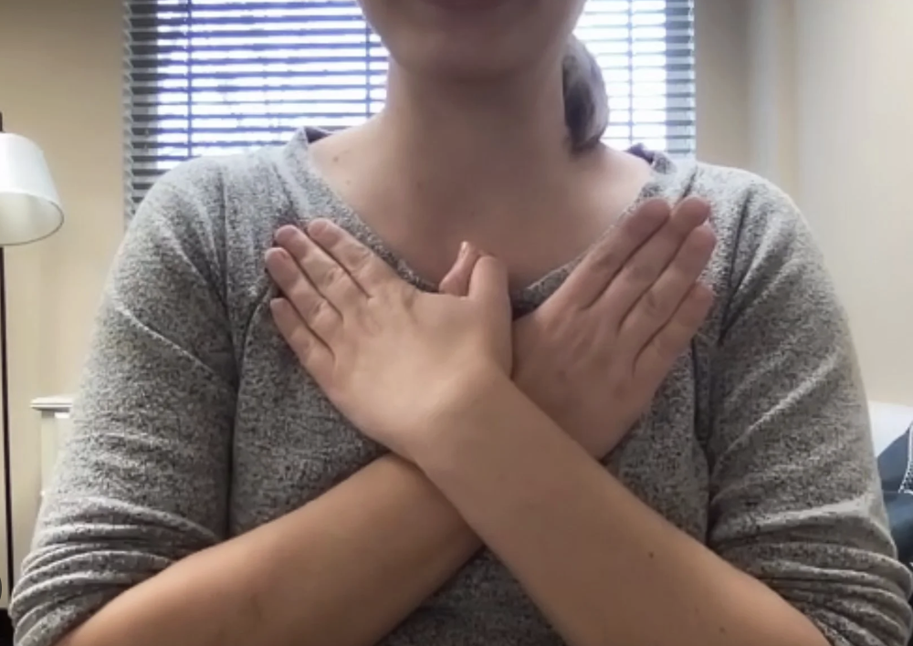 Person crossing arms over chest, standing in a room with a window with blinds behind them.