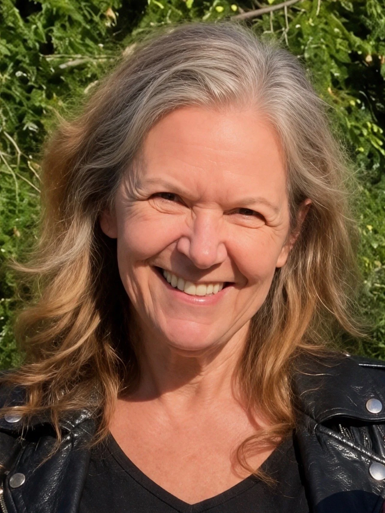 A smiling woman with long wavy blonde hair wearing a black leather jacket, outdoors with green foliage in the background.