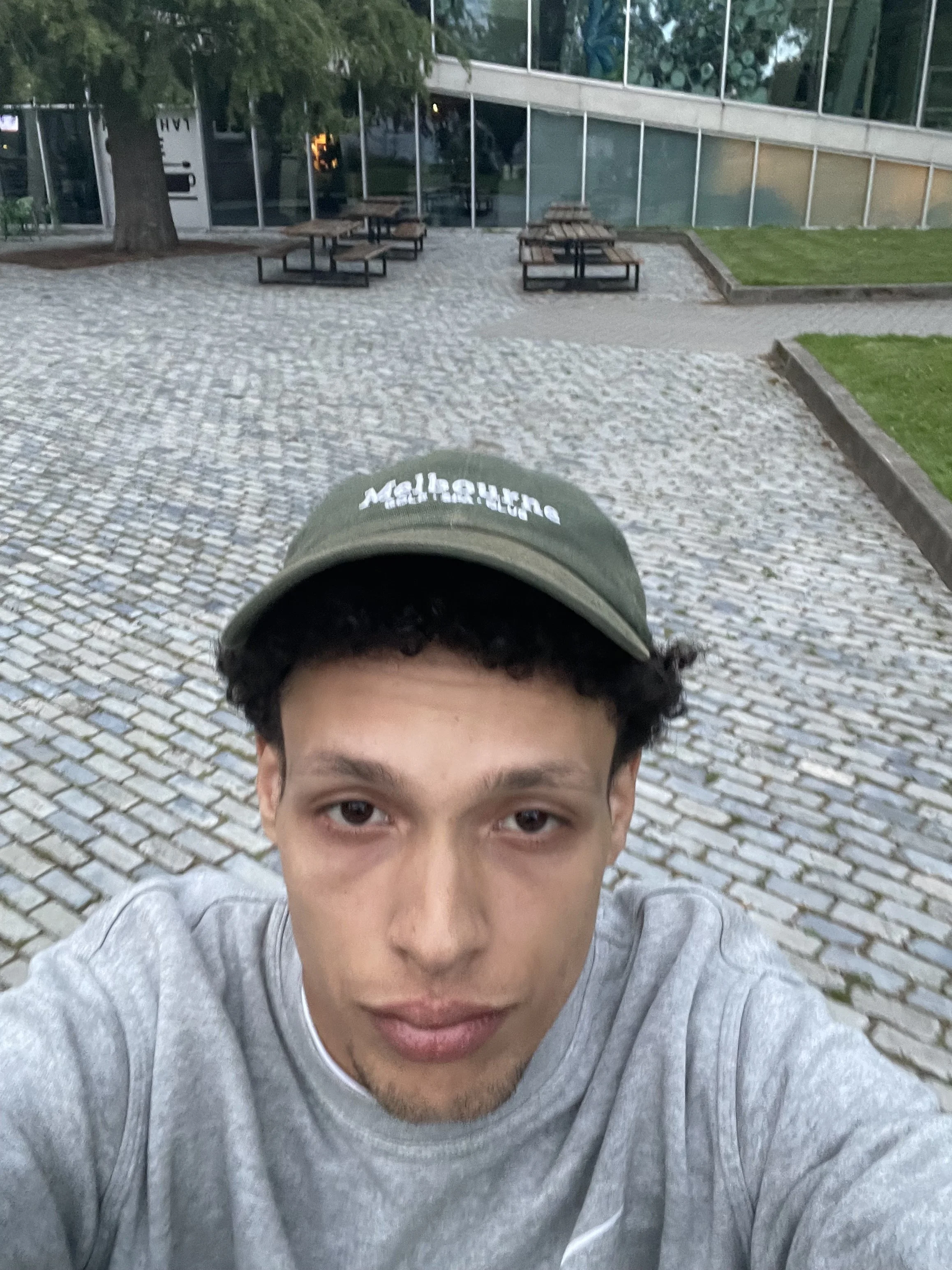 Close-up van een jonge man met een grijze trui en een groene pet, die een selfie maakt op een straat met bestraatte bestrating en buitenmeubilair, met een modern gebouw op de achtergrond.