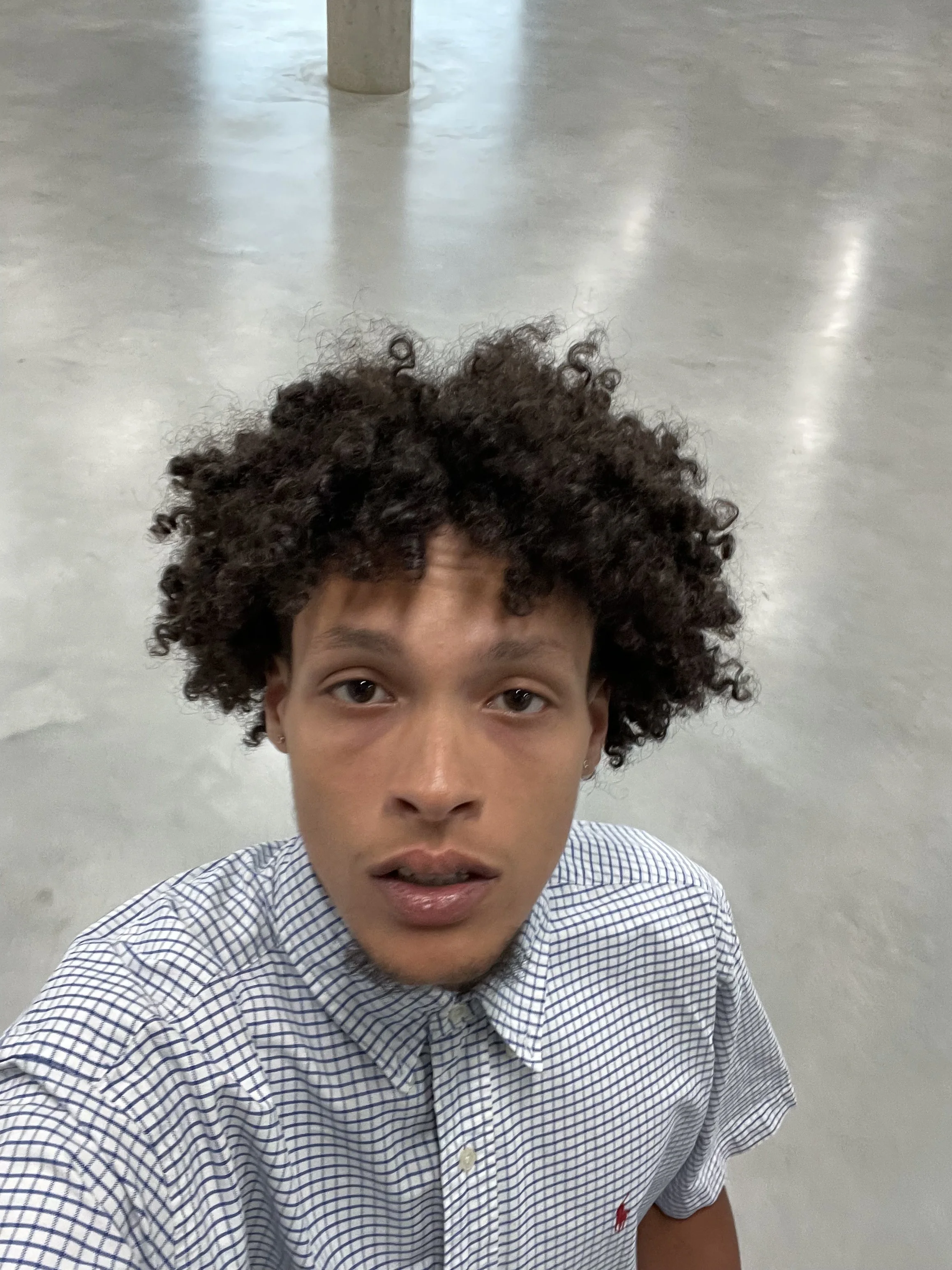 Een jonge man met krullend haar die een geruite overhemd draagt, kijkt omhoog naar de camera en staat op een lege, lichte vloer.