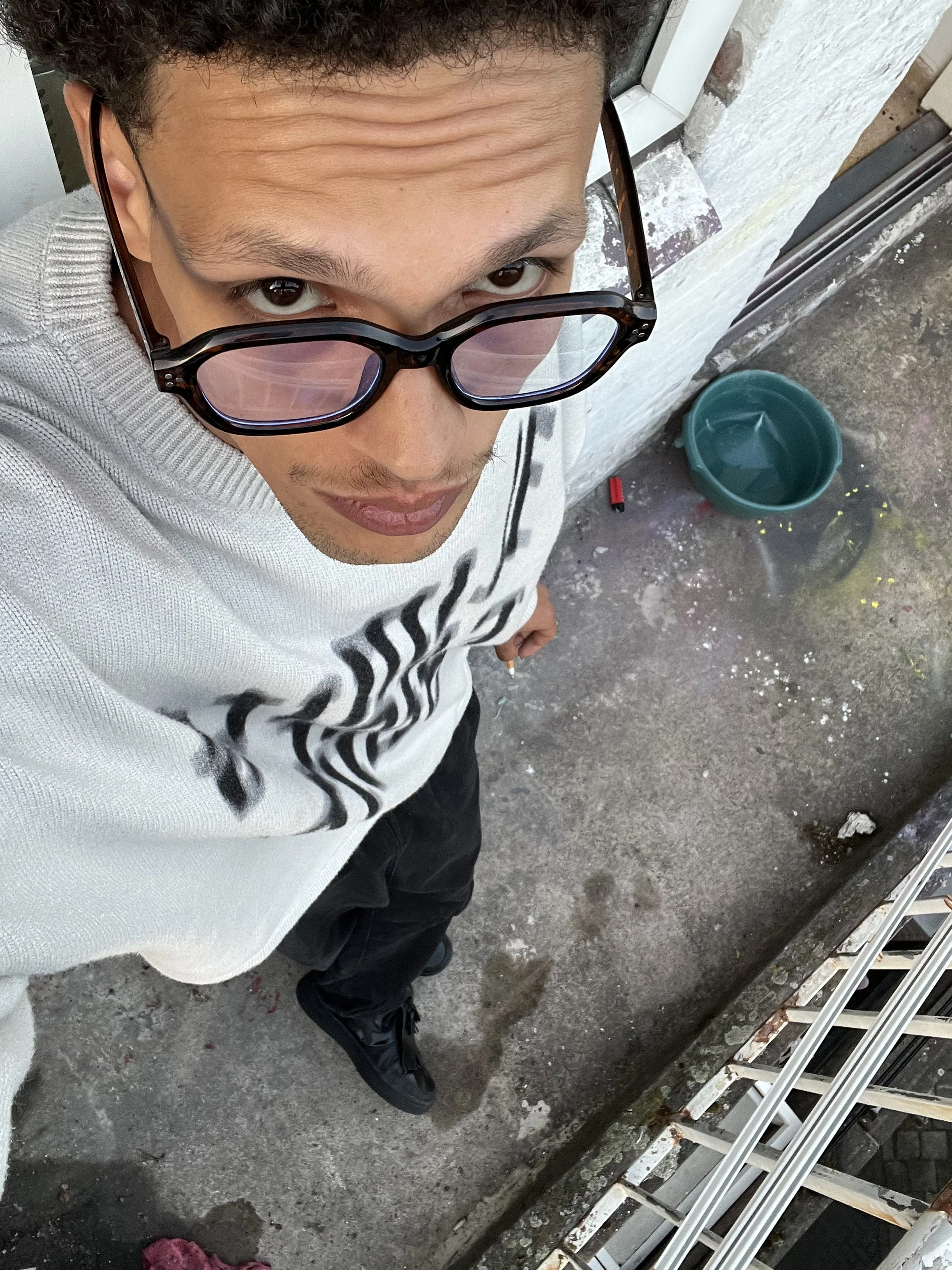 Man met zonnebril en witte sweatshirt op een terrazzo vloer, met een groene emmer en een rode lucifer in de buurt.