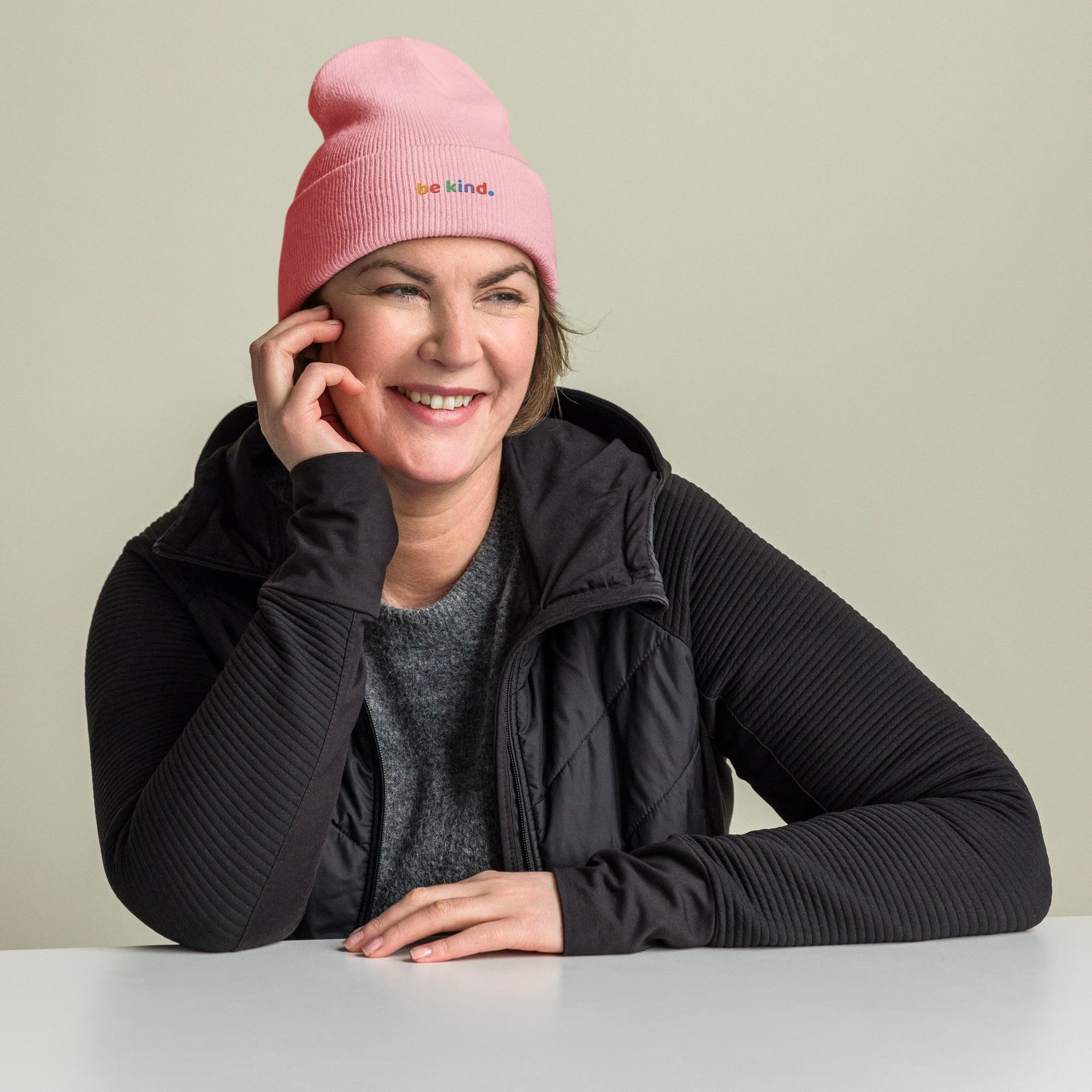 Woman in a black jacket smiling in a baby pink be kind cuffed beanie