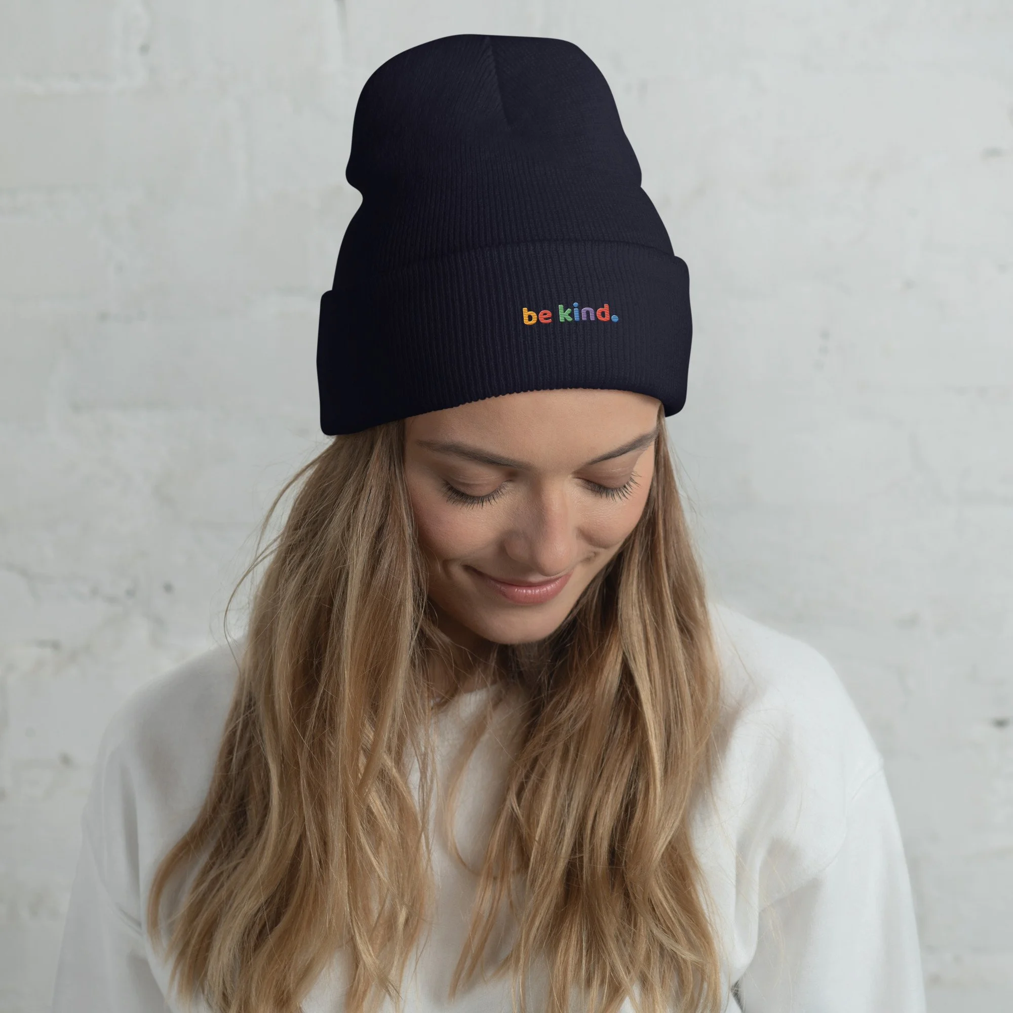 Woman with blonde hair looking down in a navy be kind cuffed beanie against a white brick wall