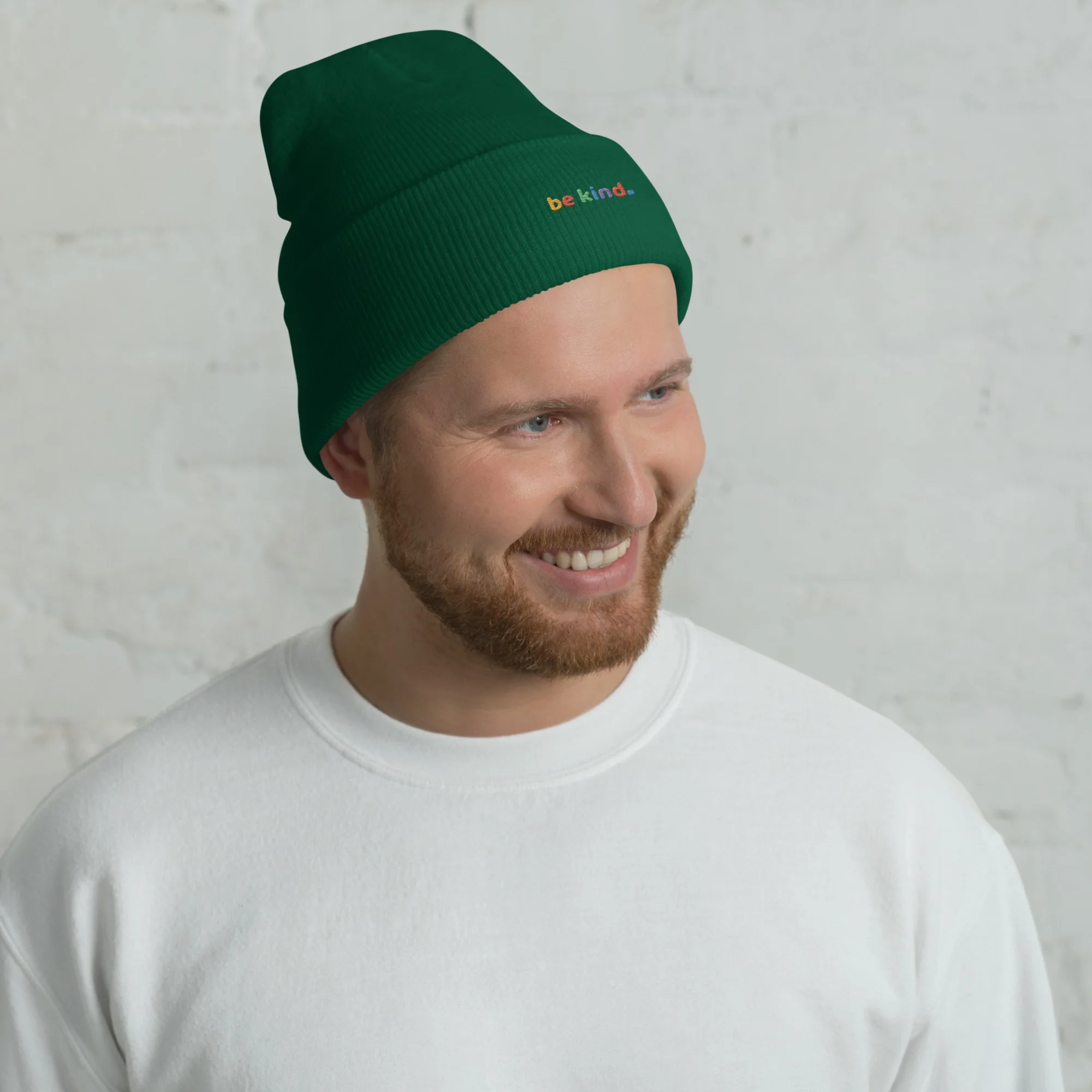 Smiling man in a white sweatshirt wearing a spruce green be kind cuffed beanie