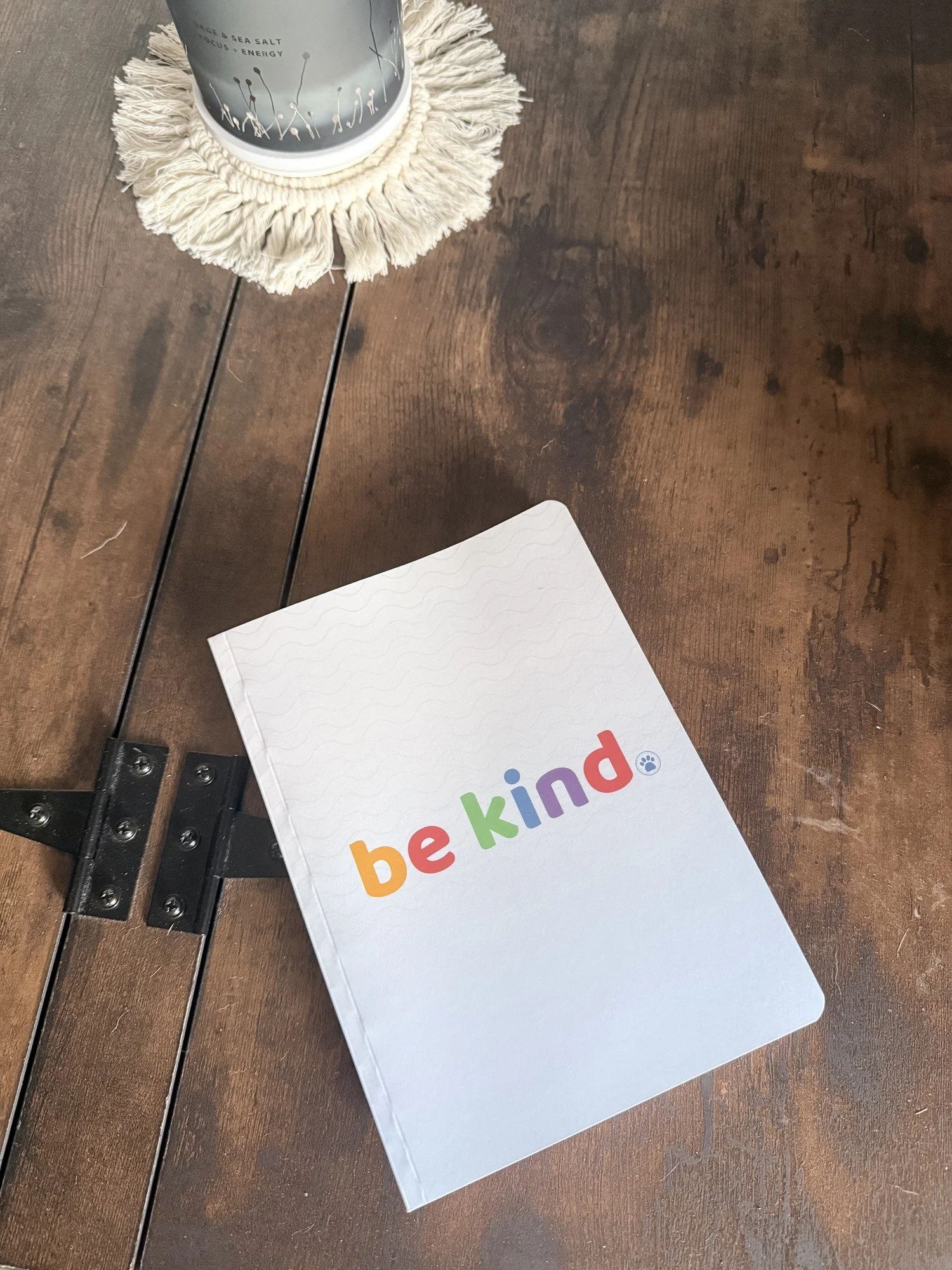 Be Kind softcover journal on a wood table, rainbow "be kind" wordmark on a white wavy-pattern cover