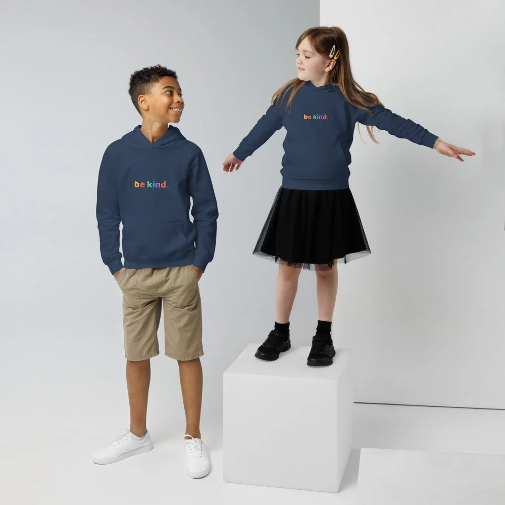 Boy and girl in navy be kind hoodies, the girl standing on a white box with her arms out