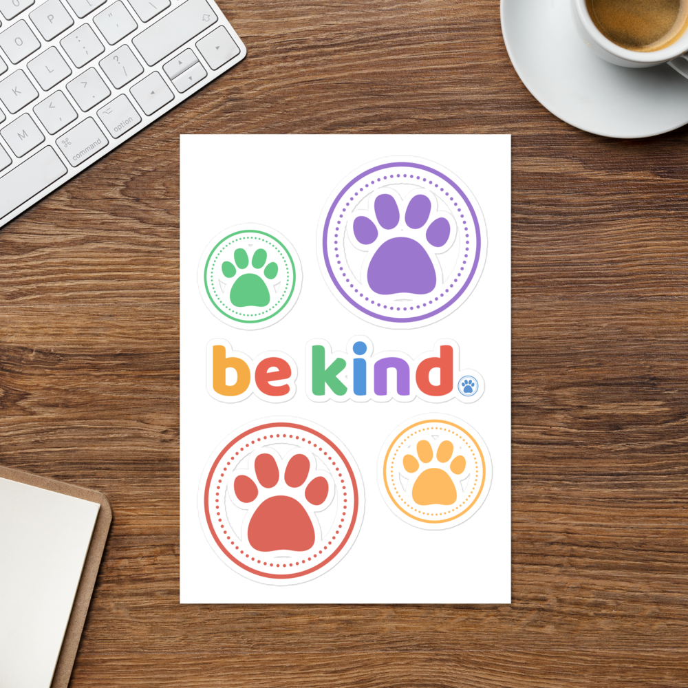 The be kind sticker sheet on a wooden desk beside a keyboard and a cup of coffee