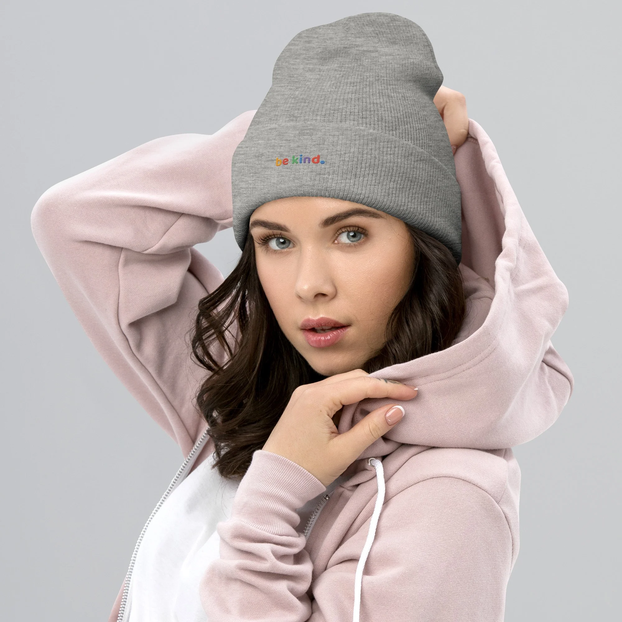 Woman in a pink hoodie pulling on a heather grey be kind cuffed beanie