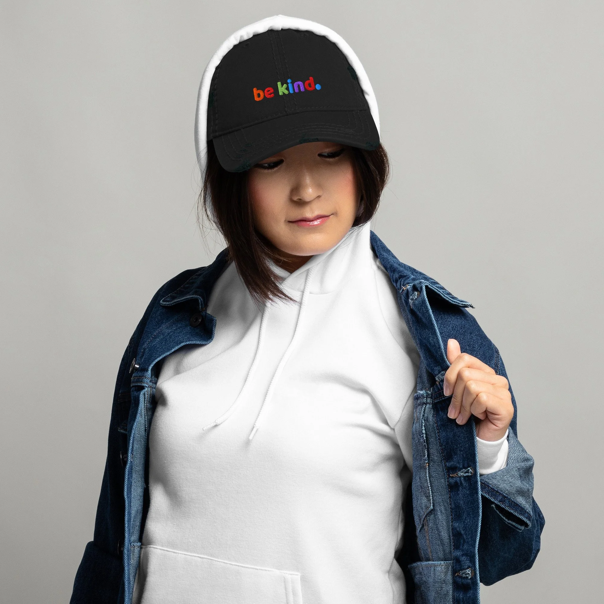 Woman in a white hoodie sweatshirt and jean jacket wearing a black distressed dad hat with be kind embroidered across the front in rainbow thread