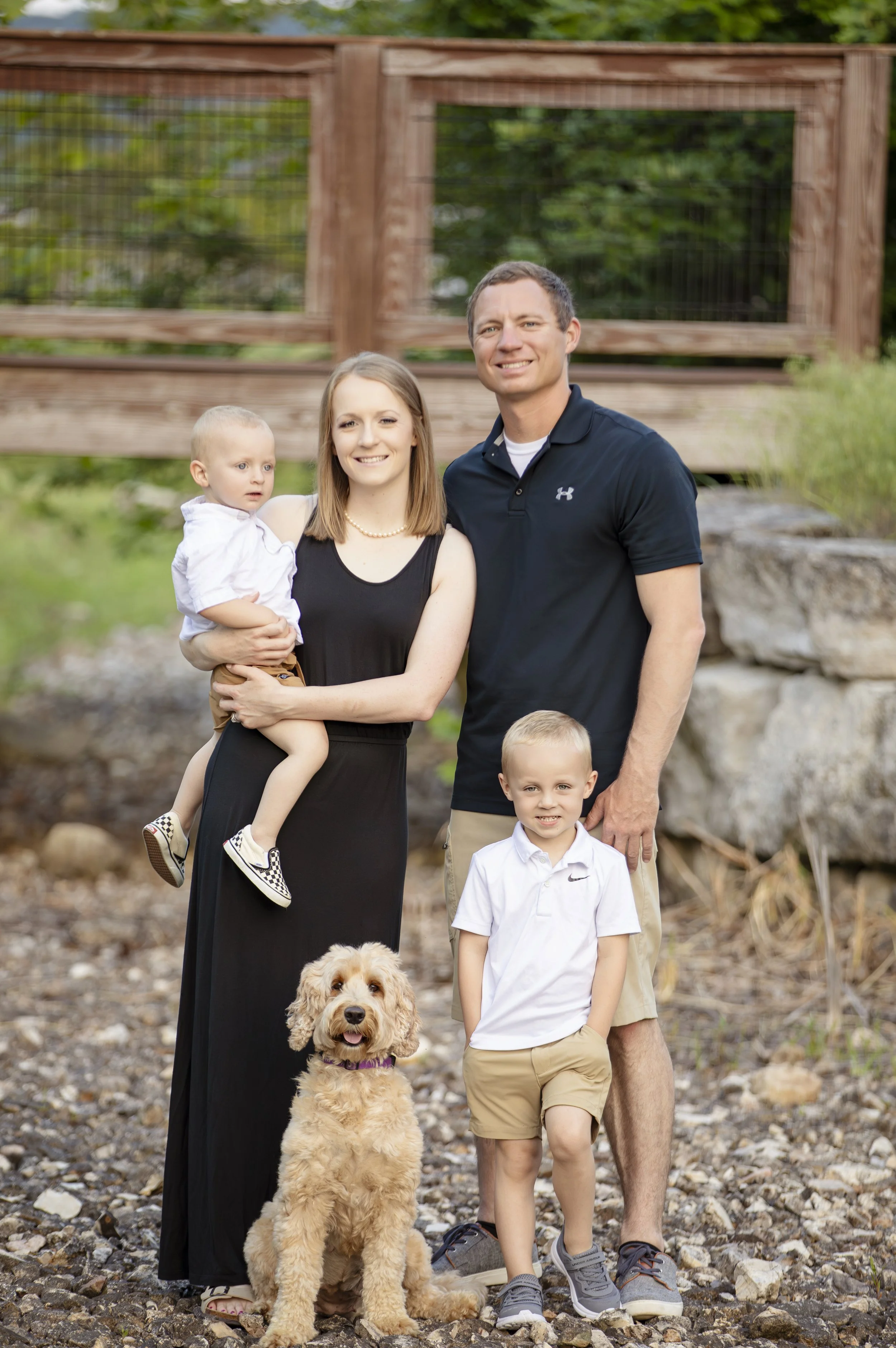 A family of five, including two adults, two children, and a dog, posing outdoors on a rocky surface in front of a wooden bridge with green foliage behind them. The woman is holding a young child, and there is a dog sitting in front of the family.