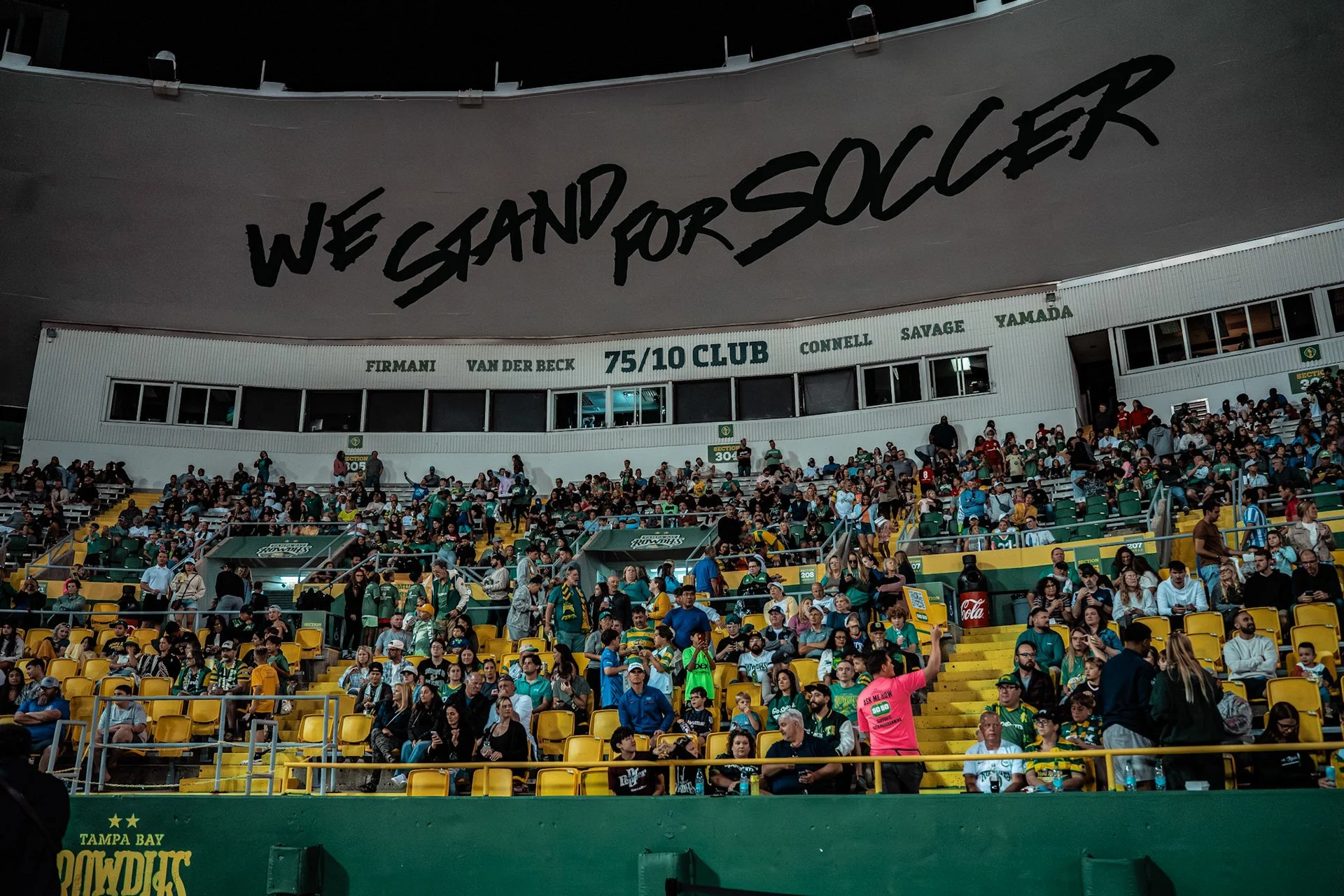 Crowd of spectators at a sports stadium, with many wearing green and yellow; large banner overhead reads 'We Stand for Soccer', indicating support for soccer fans.