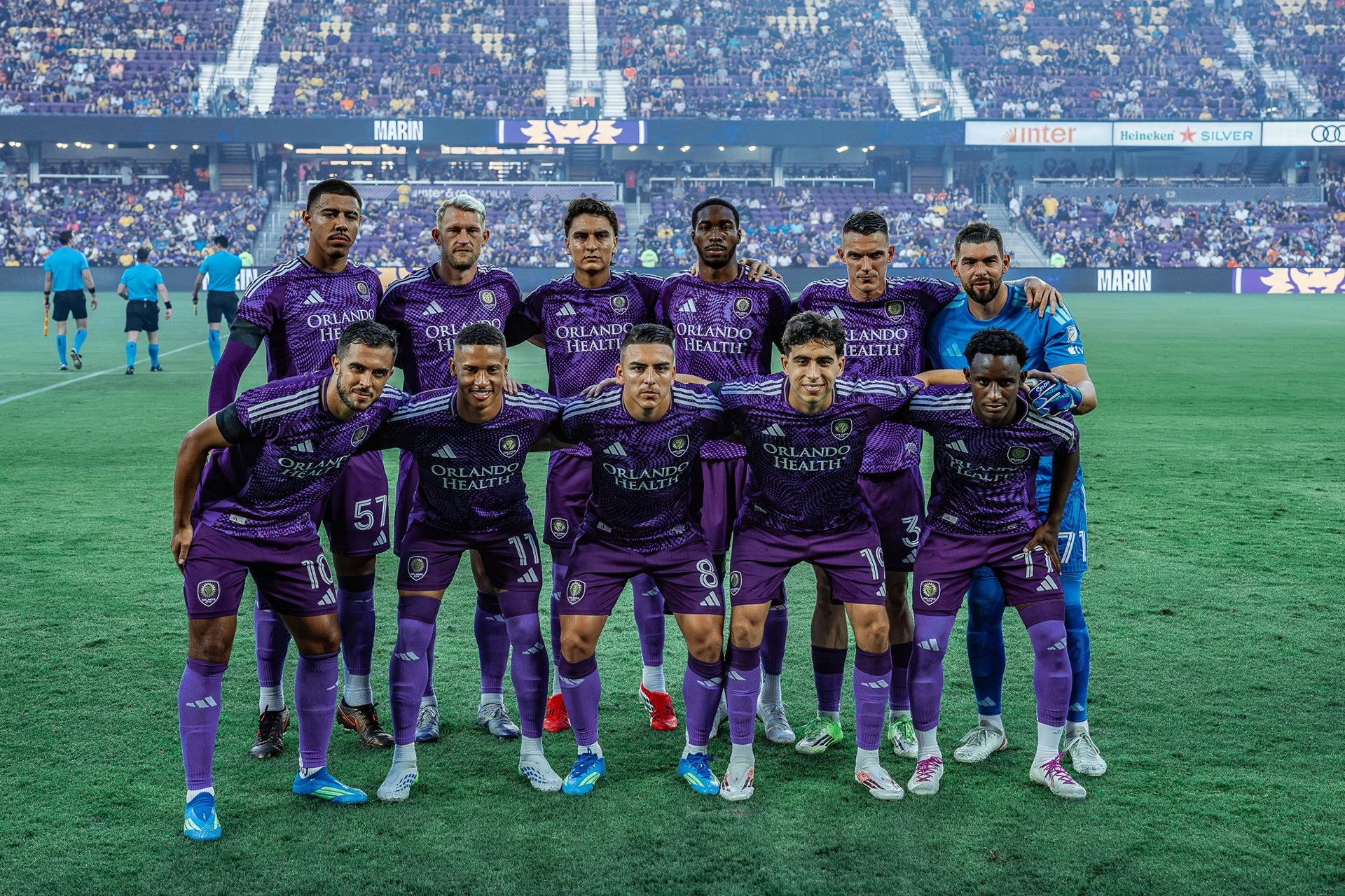 Soccer team in purple uniforms standing for a team photo on the field before a game, with stadium and spectators in the background.