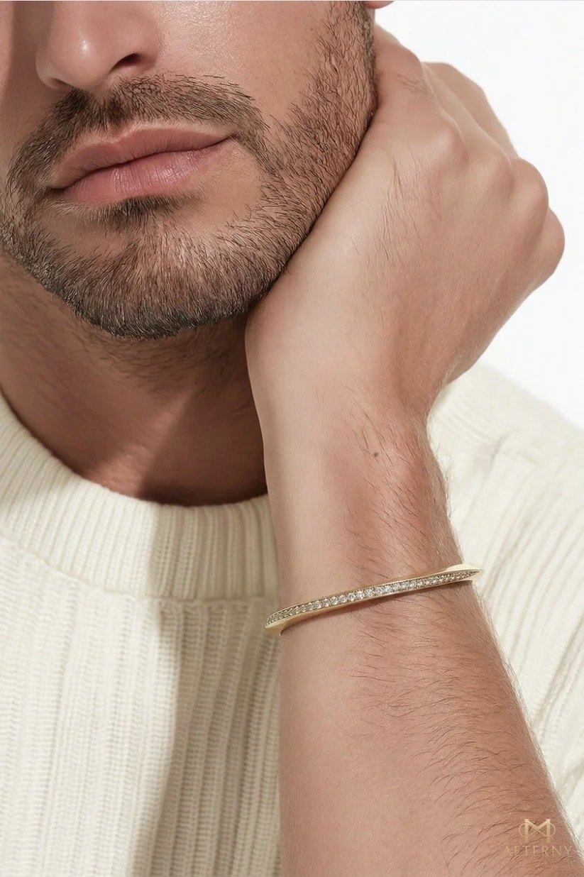 A close-up of a man's face and hand, showing his beard, lips, and nose, with his hand resting on his cheek. He is wearing a cream-colored knit sweater and a gold bracelet with small diamonds on his wrist.