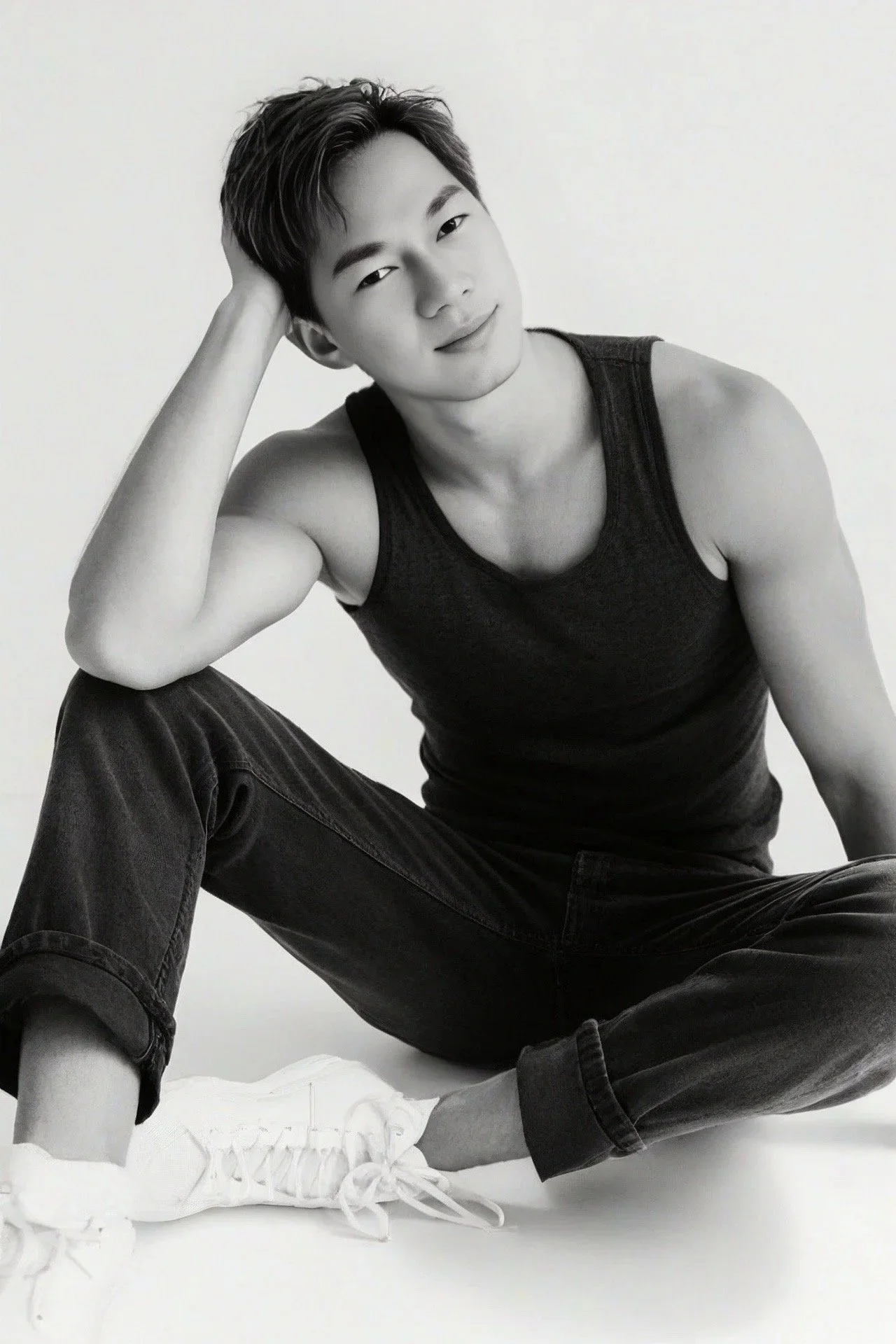 Black and white photo of a young man in a sleeveless top, sitting on the floor with one leg crossed and his hand on his head, smiling softly at the camera.