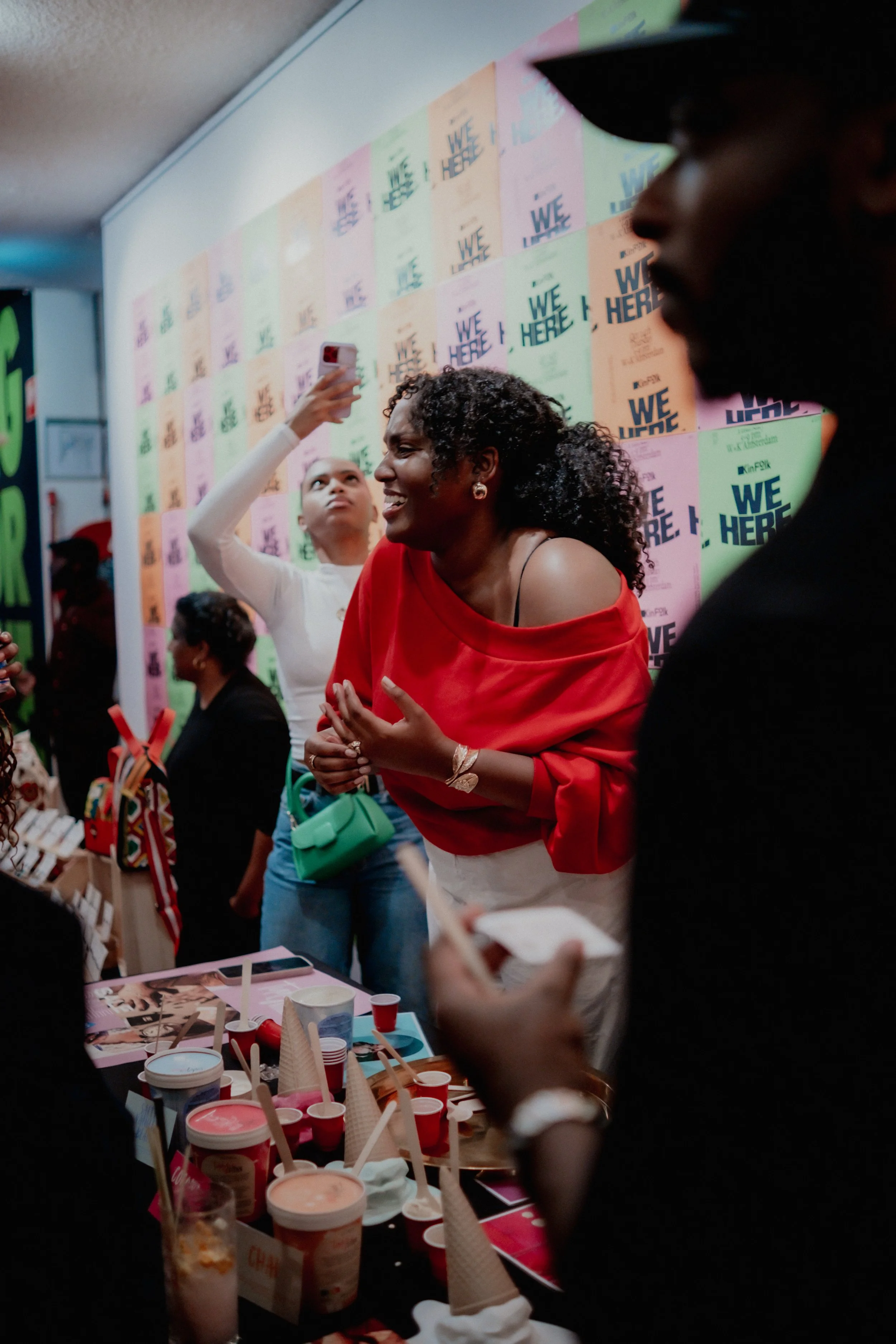 A woman in a red off-shoulder top smiling and talking to a man with a beard at an event with a colorful background and tables with ice cream, cups, and straws.