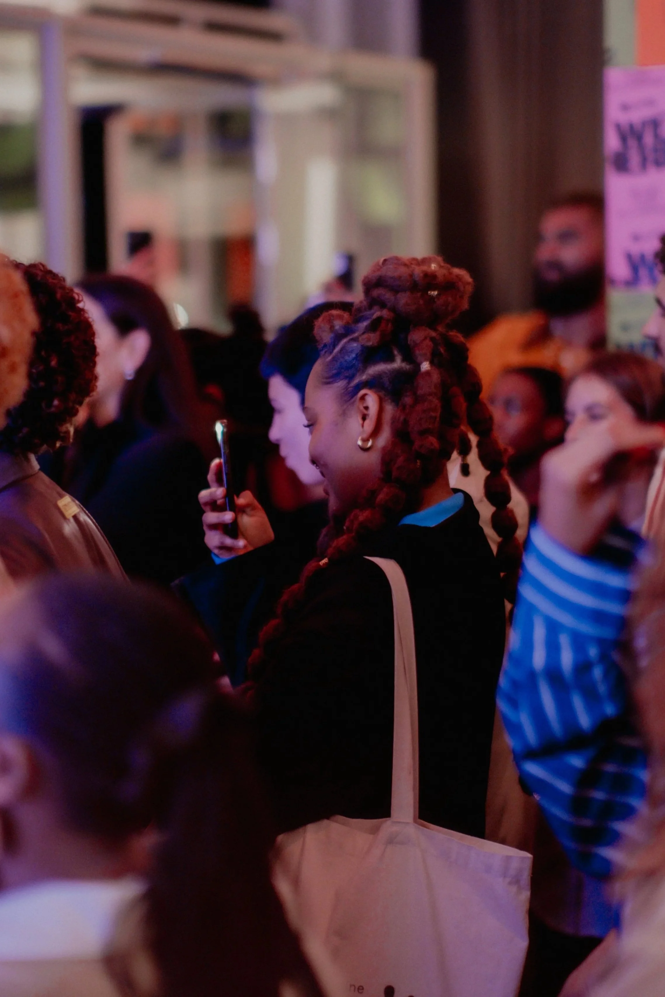 A woman with dreadlocks styled in an updo, wearing a blue shirt and earrings, is taking a video on her phone in a crowded indoor setting with dim lighting.
