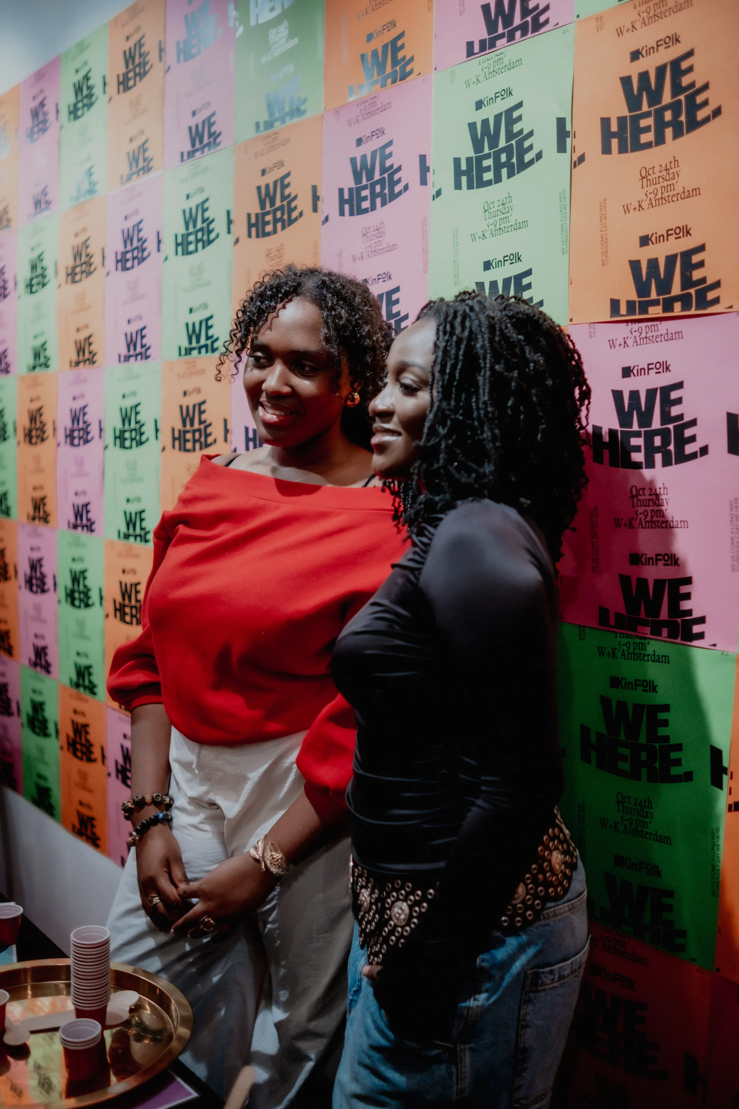 Two women stand together in front of a wall decorated with colorful posters that read 'WE HERE' and provide event details. They are smiling and posing for the photo, one wearing a red top and white pants, the other in a black top and blue jeans.