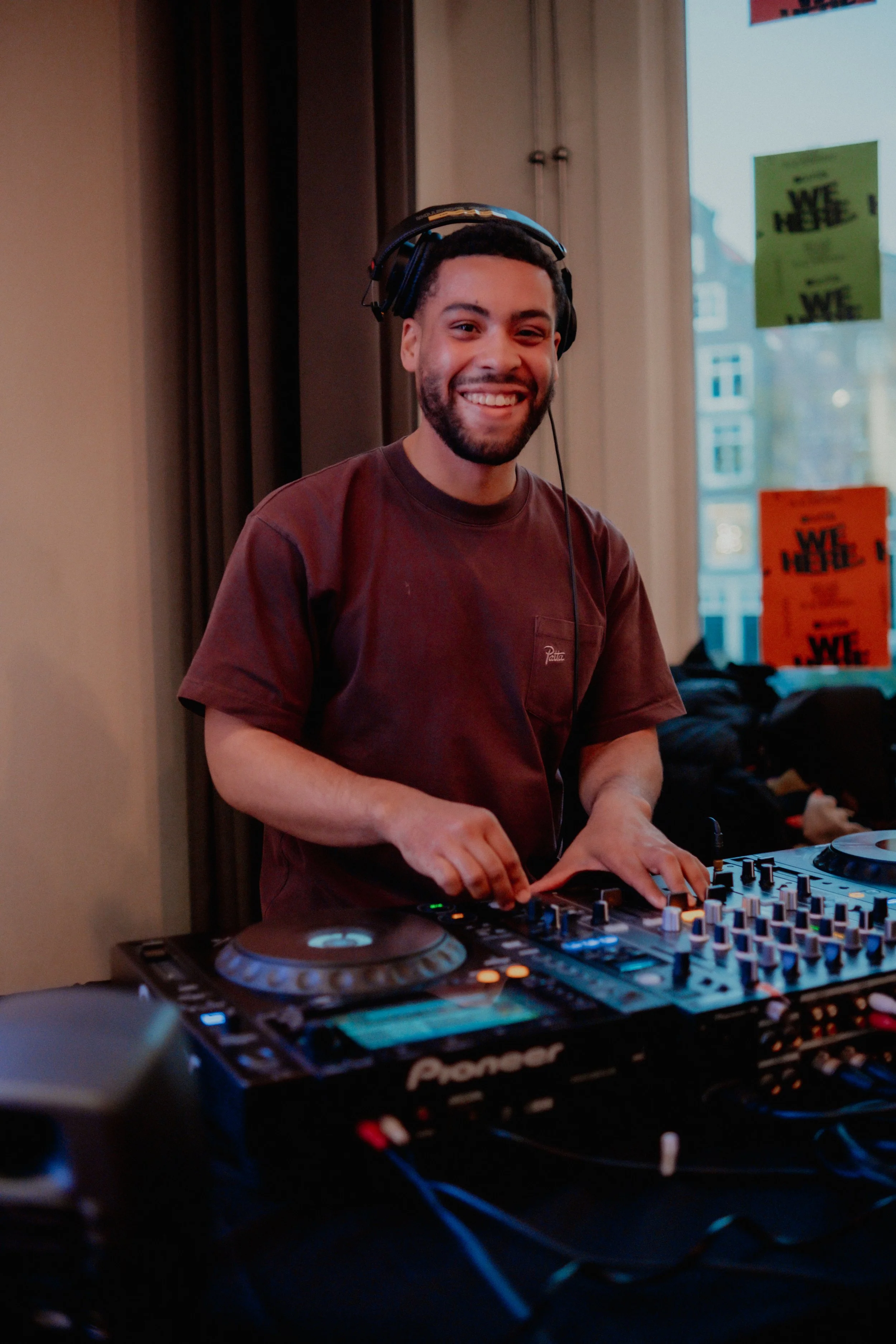 Smiling young man with a beard, wearing headphones, DJing with a Pioneer DJ controller in an indoor setting with posters and windows in the background.