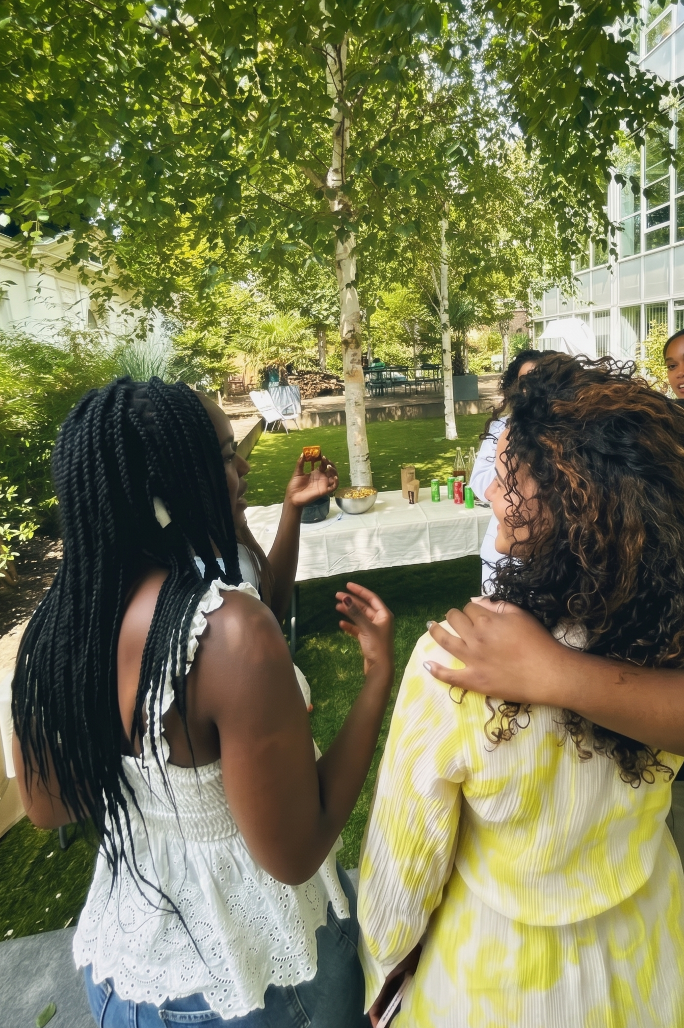 Two women are hugging outdoors on a sunny day, with a table of snacks and drinks behind them and green trees in the background.