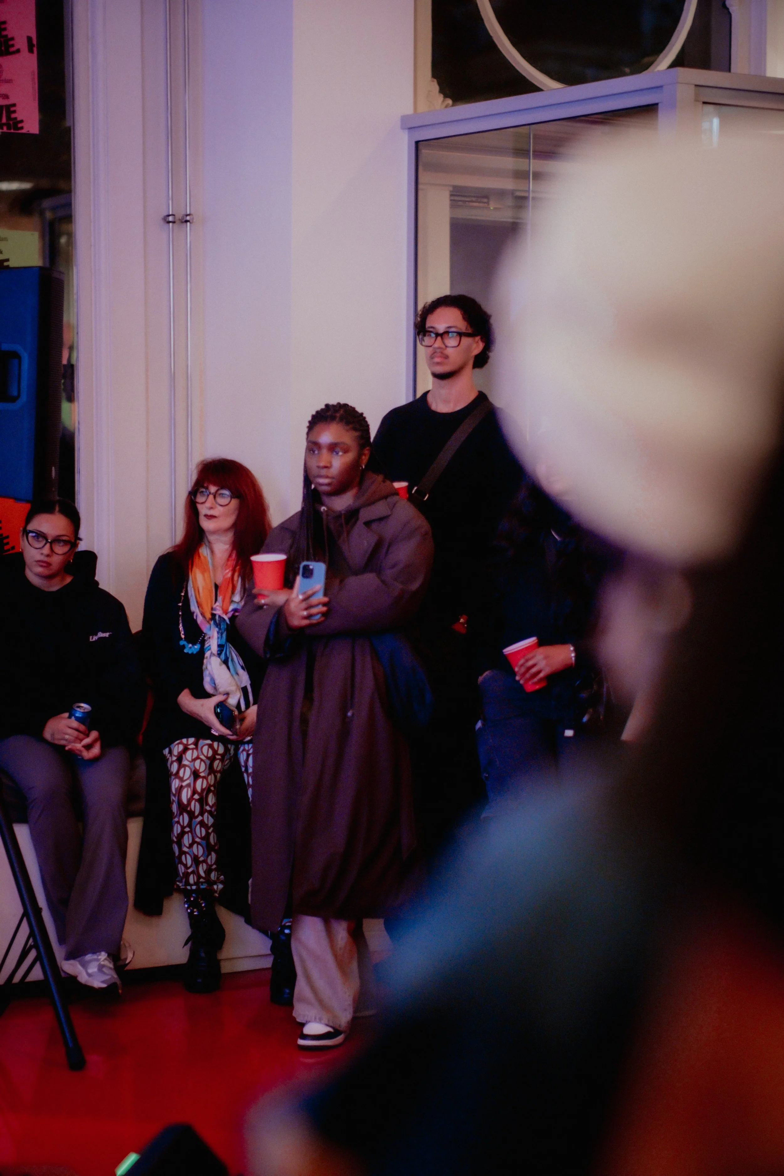 Group of people standing and sitting indoors, some holding red cups, with a person in a long brown coat and sneakers in the foreground, and a blurred area on the right.
