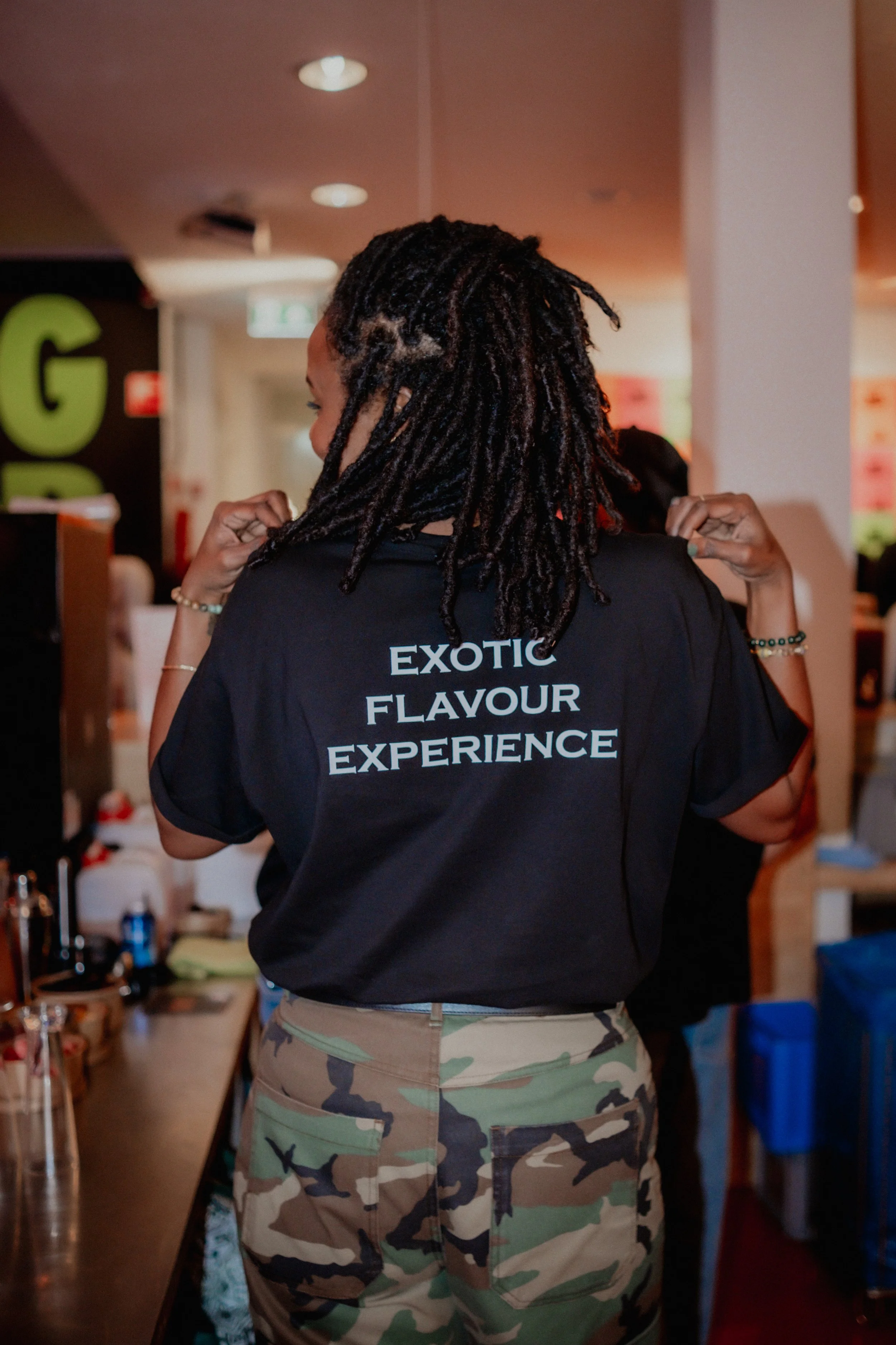 Person with dreadlocks wearing a black T-shirt and camouflage pants, standing in a room with various items on shelves.