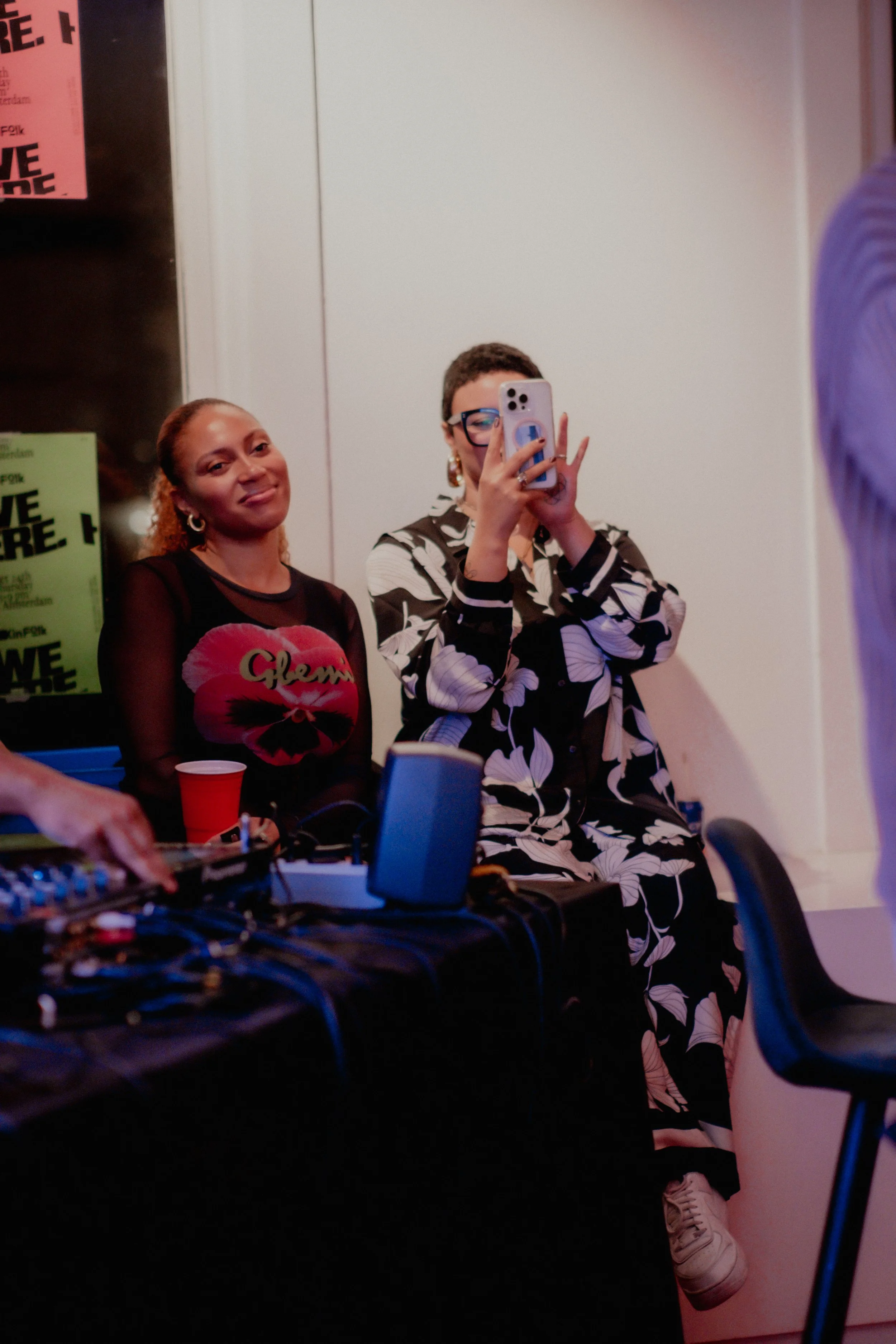 Two women sitting in front of a mirror, one is taking a selfie with a smartphone, the other is smiling. There are posters on the wall, and a DJ mixer and laptop on the table in front of them.