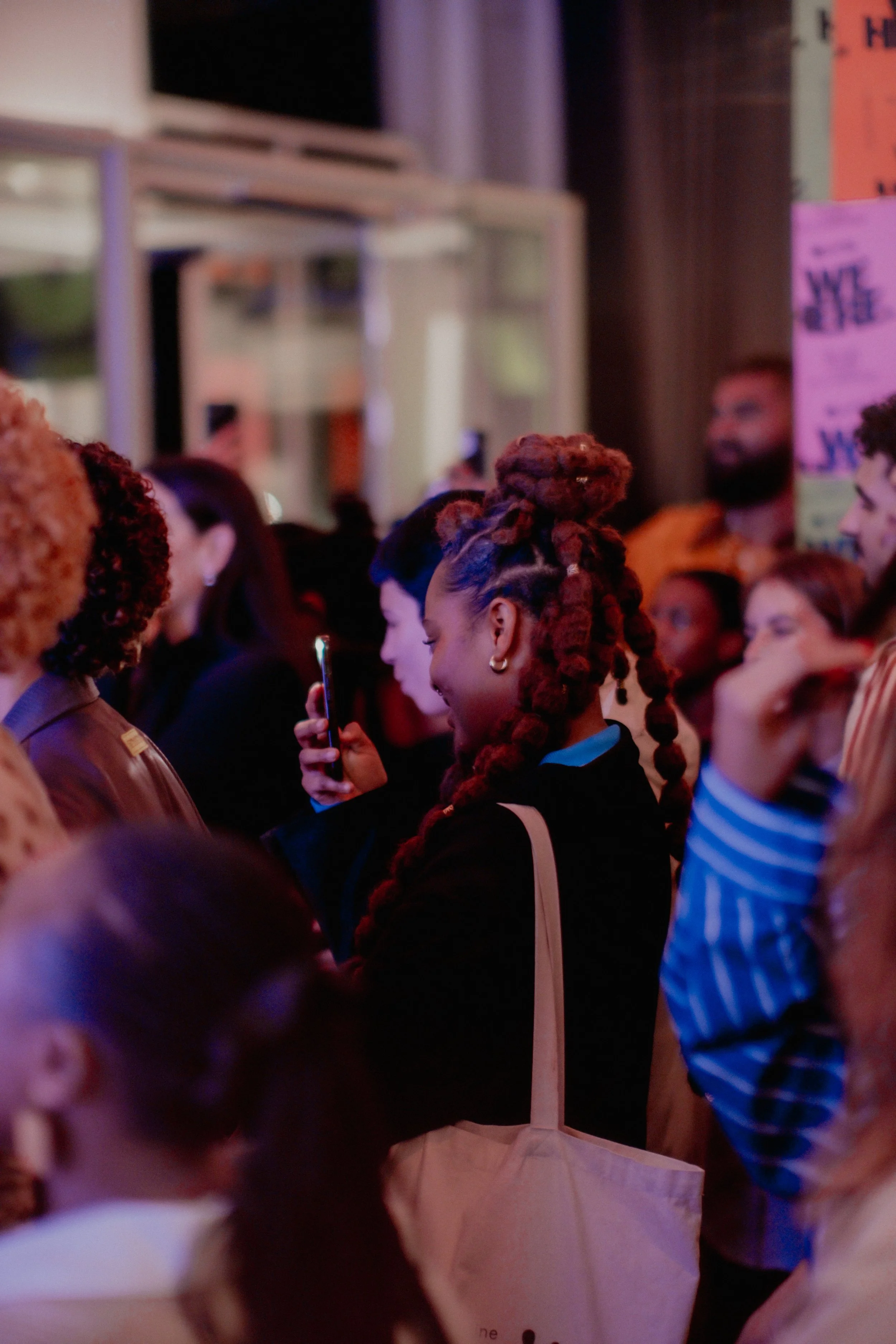 A crowd of people gathered indoors, with some focusing on a phone, titled and featuring a woman with red braided hair.