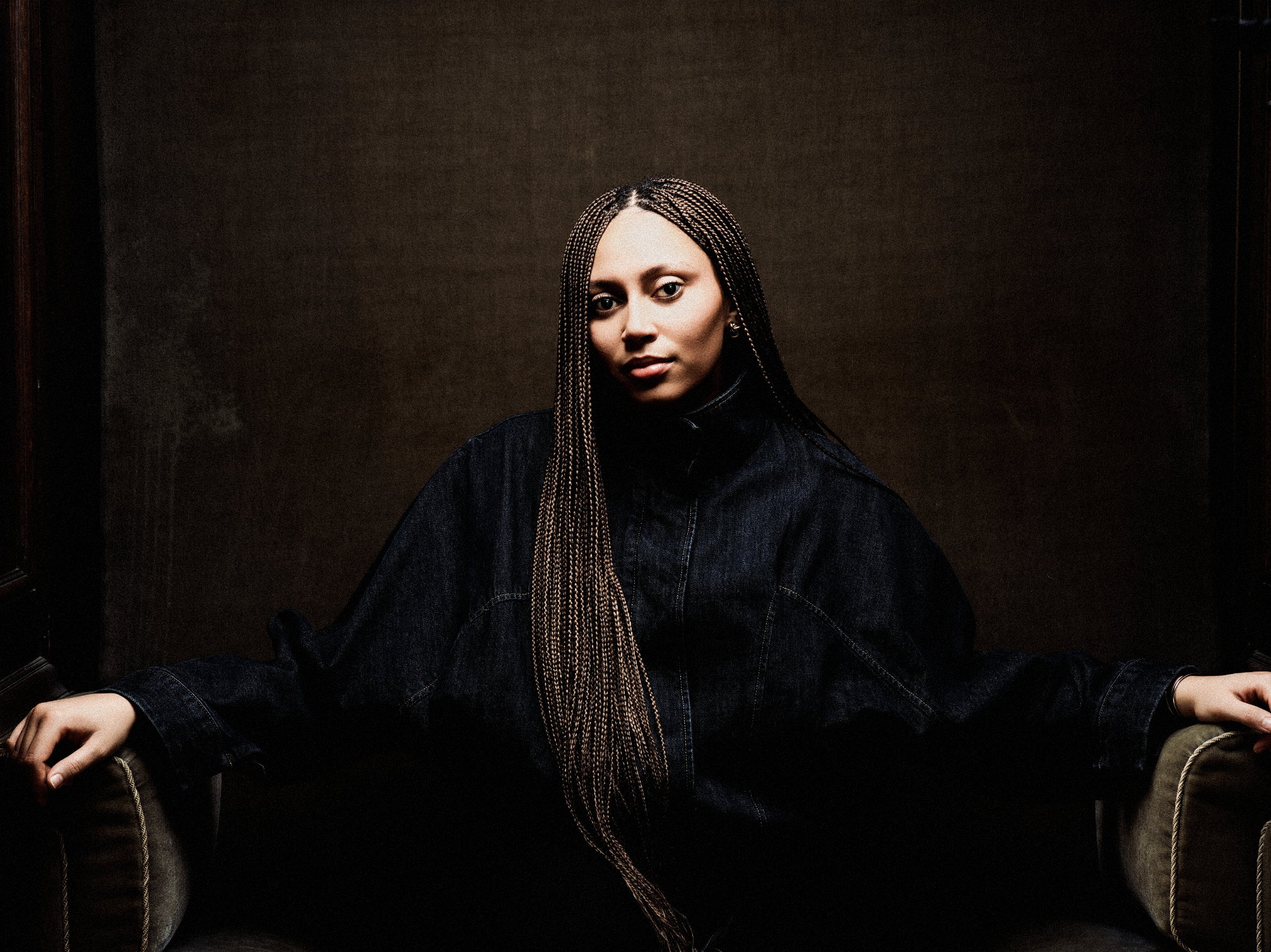 Portrait of a woman with braided hair, wearing a dark denim jacket, sitting in dark brown armchair, with a plain brown background.