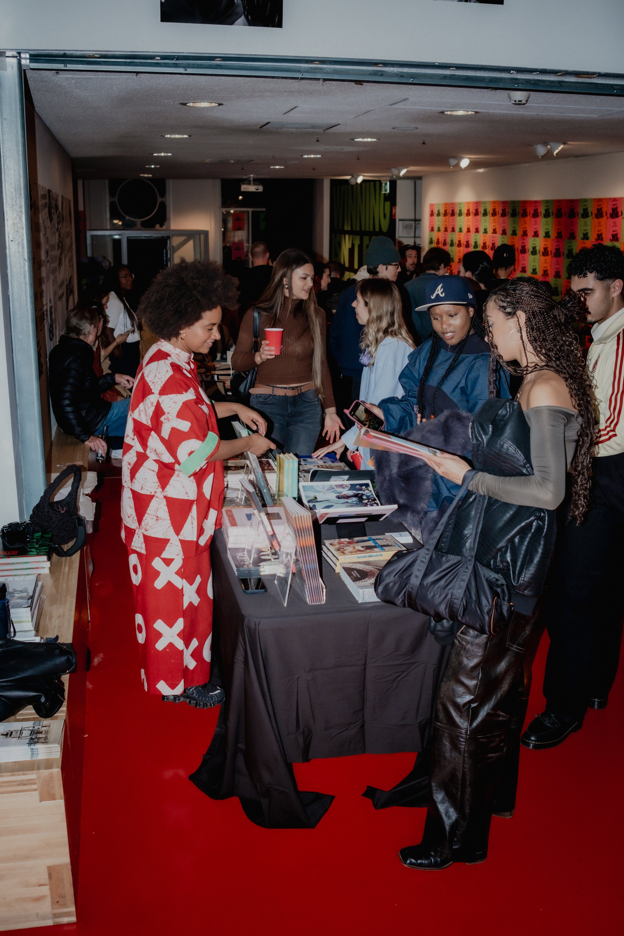People gather at a table filled with books and magazines at a promotional or book event. Some are reading, others are chatting or browsing. The setting appears to be an indoor venue with colorful decorations and posters on the wall.