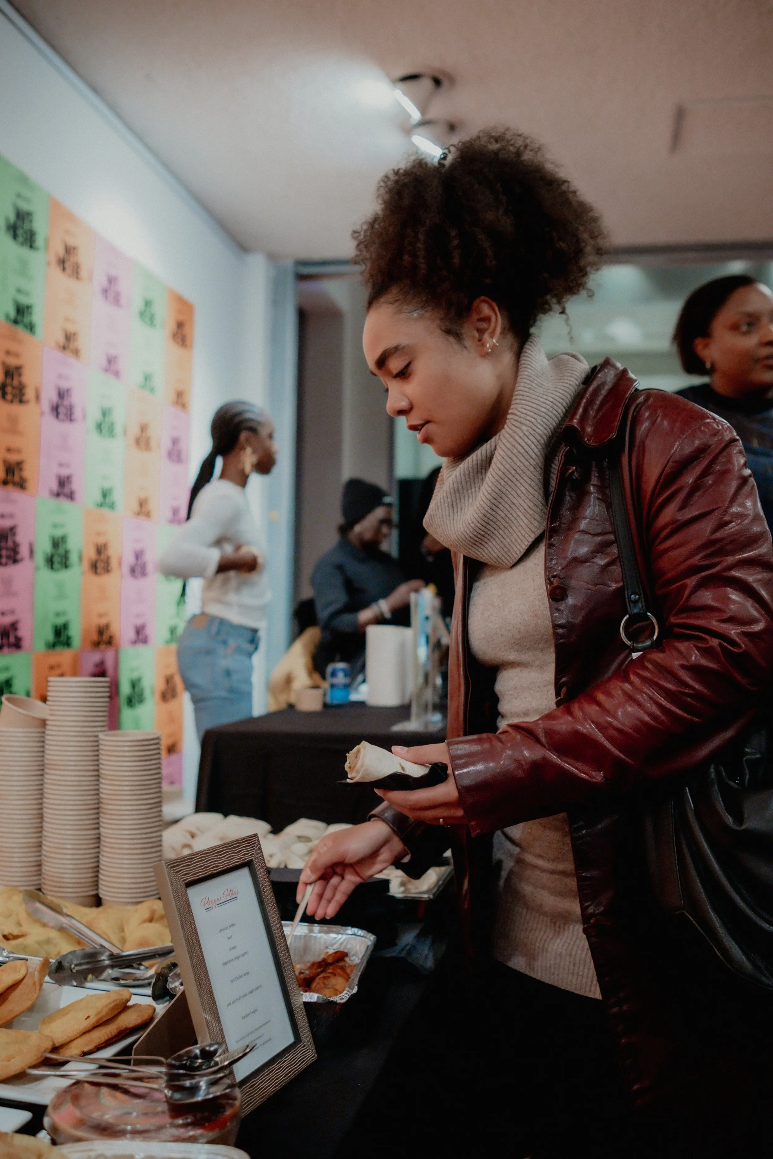 A woman with curly hair in a bun wearing a beige sweater and a maroon leather jacket is holding a wrap or sandwich at a food buffet table. She appears to be serving herself or ordering food. Other people are visible in the background, including a wom